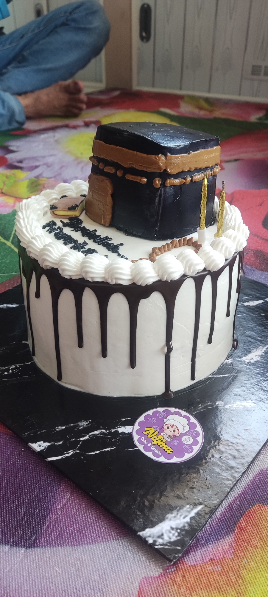 A black and white drip cake with a Kaaba replica on top is displayed on a marble cake board. The cake is adorned with white frosting piping around the top edge, and chocolate ganache dripping down the sides. Two gold candles are placed on the top layer of the cake. A small book decoration is situated to the left of the Kaaba replica. Black Arabic calligraphy is written on the cake's white frosting. A circular sticker with the text "Najma Cake & Cookies" is placed on the cake board. In the background, a person wearing blue jeans is sitting on a colorful patterned rug. The scene appears to be indoors, with a wooden door visible in the background. The lighting suggests an indoor setting.