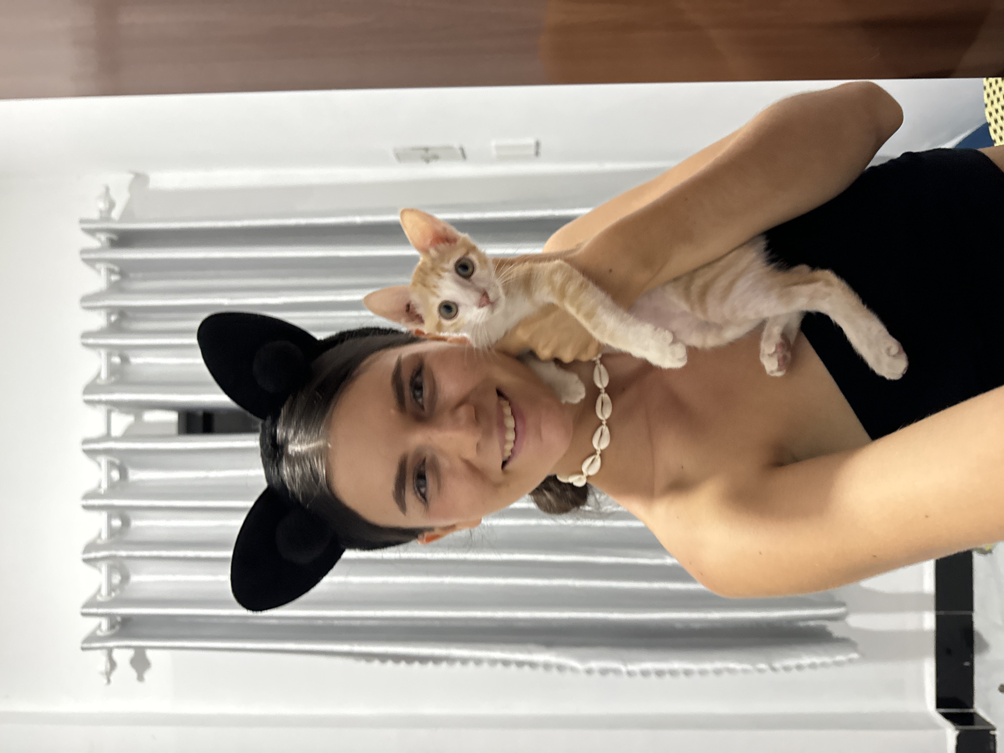 A woman wearing black cat ears and a seashell necklace holds a small, ginger and white kitten in an indoor setting. The woman is smiling and looking at the camera. She is wearing a black strapless top. The kitten is looking to the left of the frame. Behind them, a silver, pleated curtain hangs from a window or wall. The room appears to be a domestic interior in Mũi Né, Vietnam.