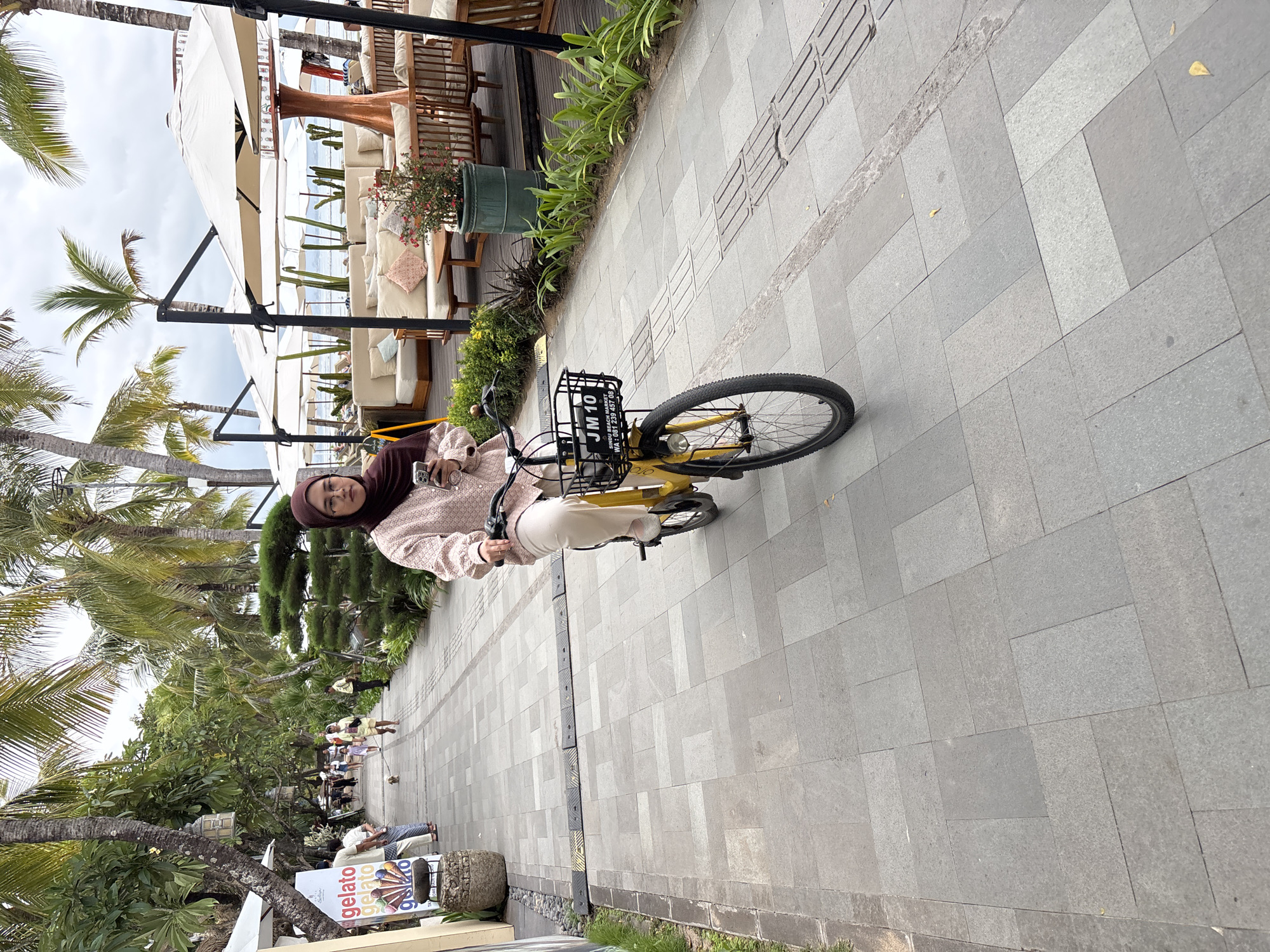 A woman wearing a dark brown hijab and a light pink long-sleeved blouse is riding a yellow bicycle down a paved walkway. She is holding a smartphone in her right hand and looking at it. The bicycle has a black basket with a sign that reads "JM 10 SINDU BEACH MARKET WA: 081 239 457 08". She is wearing light-colored pants and white shoes.
The walkway is lined with lush green trees and palm trees. In the background, there are outdoor seating areas with white umbrellas, couches, and tables, suggestive of a cafe or restaurant, possibly by the beach. Further down the walkway, other people can be seen walking, including a person walking a small dog. The pavement is made of large, rectangular grey tiles. The overall scene depicts a lively pedestrian area in a tropical location, likely Denpasar, Indonesia, given the presence of palm trees and the style of the establishment.