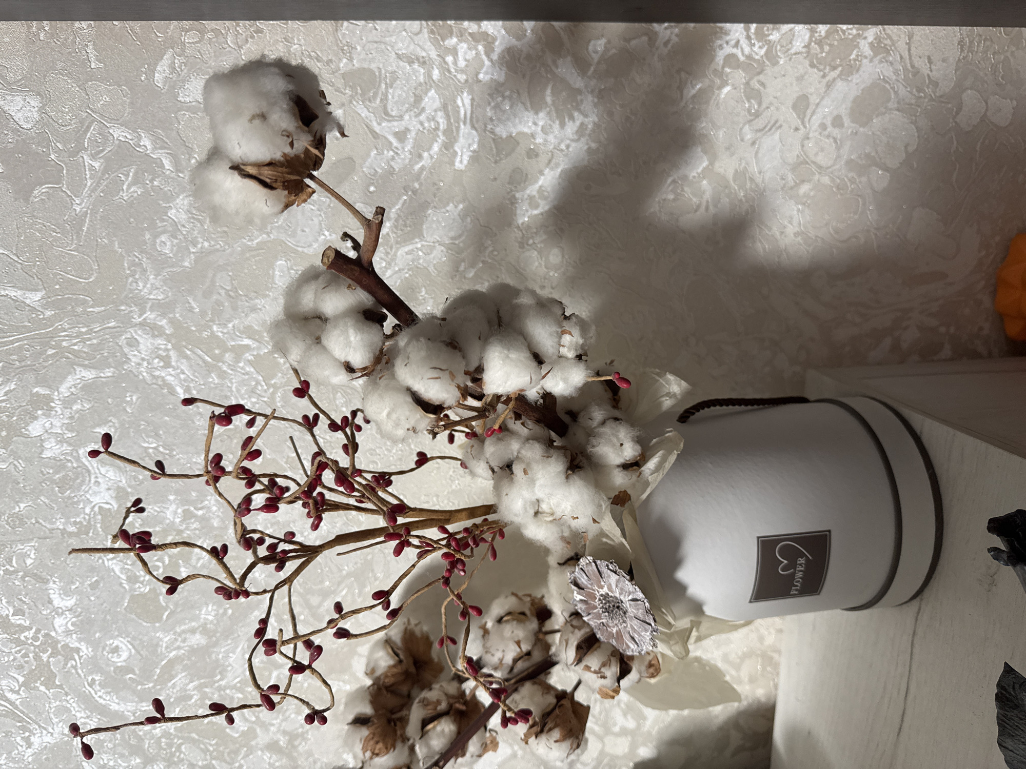 A dried floral arrangement featuring cotton bolls and red berries is displayed in a white round box. The box has a brown trim and a square label with a heart and the word "FLOWER" on it. The arrangement is set against a textured, light-colored wall with a marbled pattern. The scene appears to be indoors, possibly on a shelf.