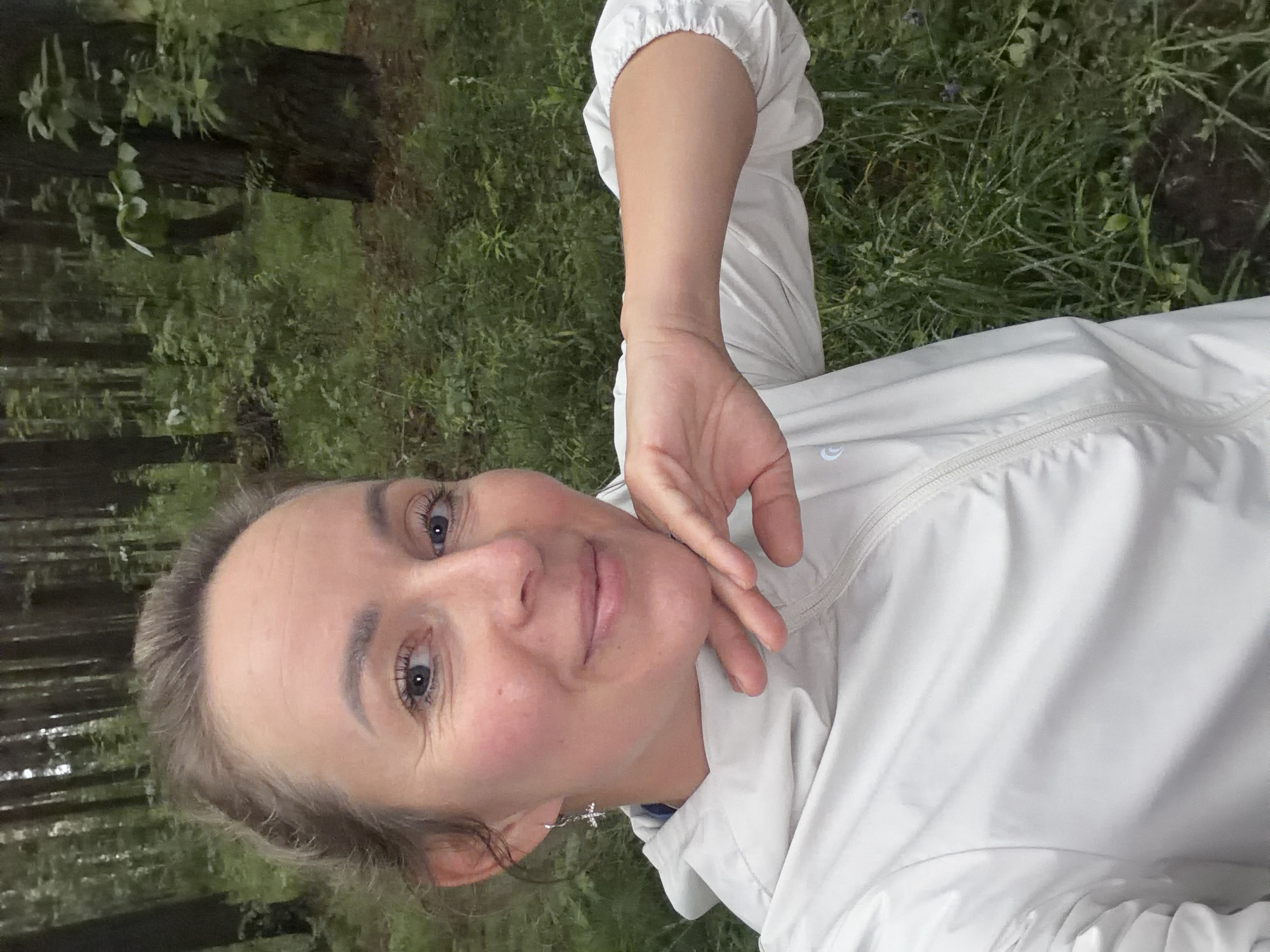 A woman is taking a selfie in a forest. She is wearing a light-colored hooded jacket. Her right arm is bent at the elbow and her hand is resting on her chin. She is smiling and looking at the camera. The background is a forest with trees and green foliage. The lighting appears to be overcast, suggesting it might be a cloudy day. There are no recognizable landmarks that would pinpoint this location to Chaikovsky, Russia.