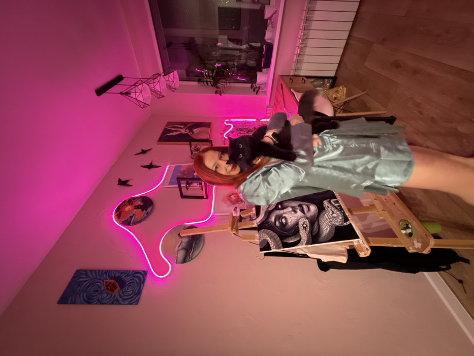 An interior scene in Rostov-on-Don, Russia, features a female subject holding a black cat, illuminated primarily by magenta neon light. The female, with reddish-orange hair, wears a light blue satin short pajama set and stands in the center-right, looking forward with a slight smile. To her left, a wooden easel displays a detailed grayscale painting of a woman's face intertwined with snakes. The easel holds a small tray with paint supplies.
On the wall behind the subject and easel, a prominent magenta neon light outlines an abstract, undulating design, wrapping around a circular painting of a dark wave and partially obscuring a vertical abstract artwork. Further right on the wall, multiple framed pictures are visible, including one featuring a white rabbit, along with three black bird-shaped wall decorations. A modern chandelier with three geometric, open-cage lamps hangs from the ceiling.
To the right, a desk with a light-colored wooden top and a matching chair is positioned beneath a window. Several potted plants are arranged on the desk and windowsill. A white radiator is located directly beneath the window, which offers a view of nighttime city lights. The room features light-colored walls and light-toned wooden flooring.