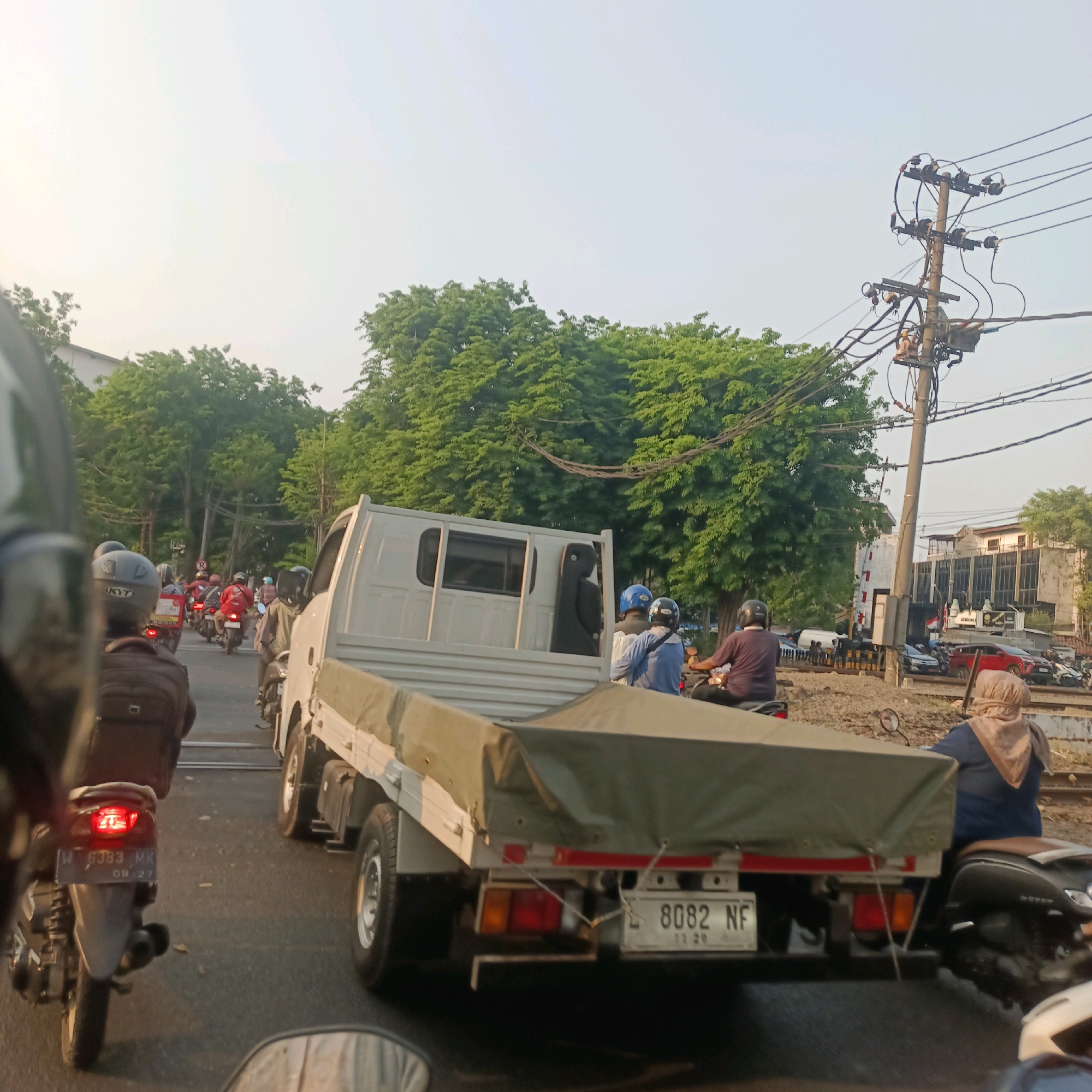 The image captures a traffic scene, likely in Surabaya, Indonesia, given the license plate formats and general urban environment. The primary focus is a white flatbed truck with a green tarp covering its cargo, stopped in traffic. The truck's license plate reads "L 8082 NF". Several motorcycles are visible, some with passengers. One motorcycle in the foreground, on the left, has a license plate "L 383 MK". The sky is bright and hazy, suggesting it might be late afternoon or early morning. Trees line the street, and utility poles with numerous wires are prominent on the right side of the image, indicating an urban setting. In the background, there are buildings and a railway crossing with barriers. People on motorcycles are wearing helmets, and some are dressed in casual clothing. The overall impression is of a typical traffic jam.