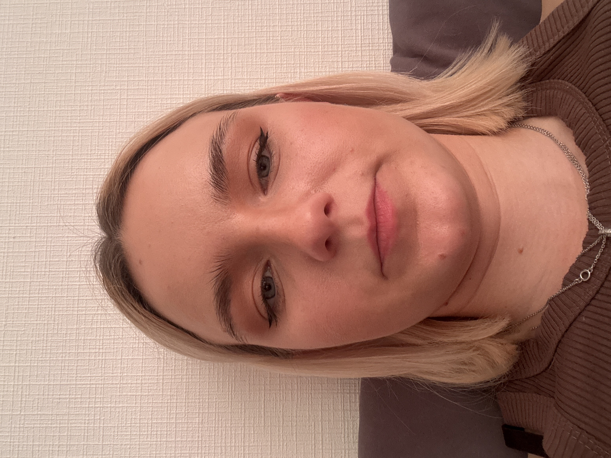 A close-up, rotated portrait shot captures a young woman from the chest up, positioned against a textured light-colored wall and a plain dark grey/brown surface, likely a pillow or blanket. Her head is tilted to her right, and the image is rotated clockwise, making her appear to be lying down.
The subject has fair skin, blonde hair parted on her left side, and light-colored eyes. Her eyebrows are full and natural. She wears visible black winged eyeliner and subtle pink lip color. A delicate silver chain necklace is visible around her neck, draped over her brown ribbed top. Small moles or freckles are present on her forehead, cheek, and neck.
Her expression is neutral, with her lips slightly parted. The lighting is soft and even, suggesting an indoor setting. There is no discernible specific activity beyond posing for the portrait. No text is visible in the image. The location is unknown, but context suggests Russia.