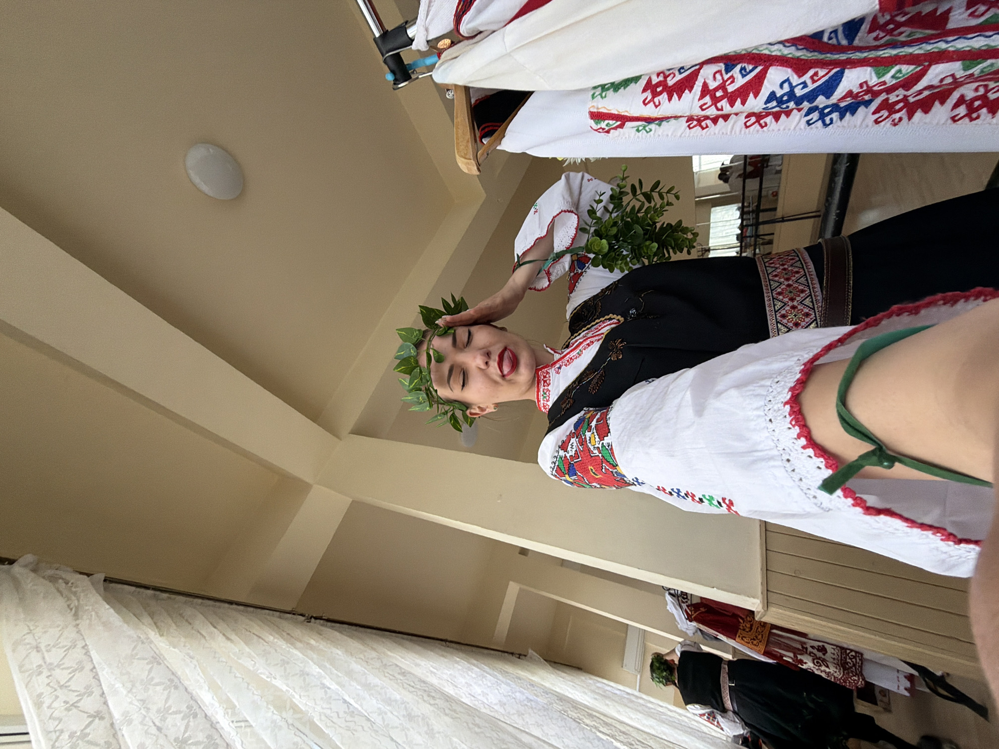 A woman in Bulgarian folk attire with a laurel wreath on her head and a bouquet of greenery in her hand stands inside a room. She is sticking her tongue out slightly, giving a playful expression. The room features neutral-toned walls, a ceiling light, and white curtains near a window. Clothes are hanging in the background. Another person in similar attire is seen farther in the background. The city of Sofia, Bulgaria is not discernable in the image.