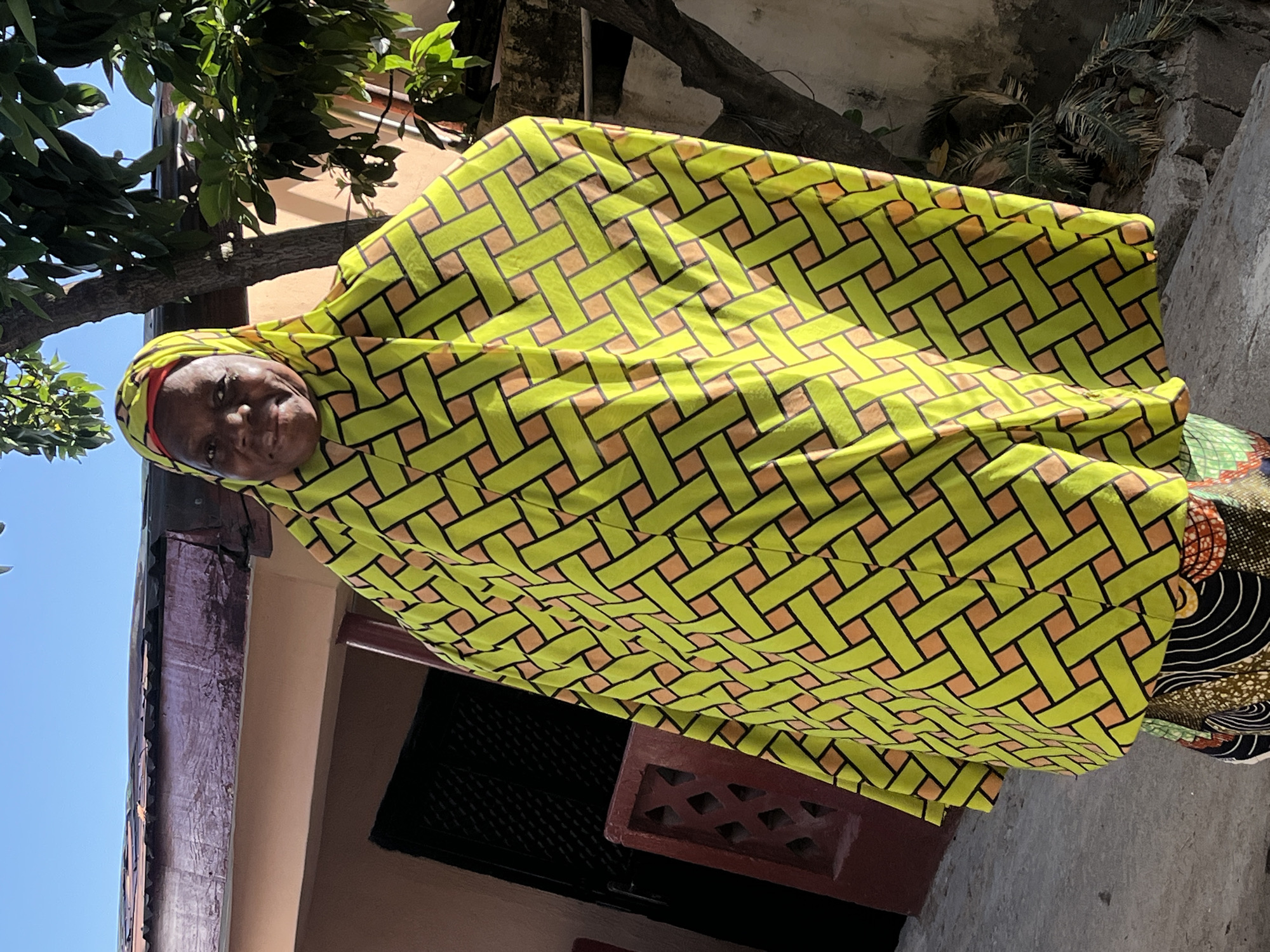 A mature woman stands outdoors, wearing a vibrant yellow and brown patterned wrap-around covering her head and body. The patterned fabric features interconnected rectangular designs in black, yellow, and brown, completely obscuring her clothes. The woman's face is visible and she appears to be looking directly at the camera.
The setting appears to be outdoors near a building with a partially visible dark-colored trim. The background includes leafy foliage and a bright blue sky. The picture was taken in Jos, Nigeria.