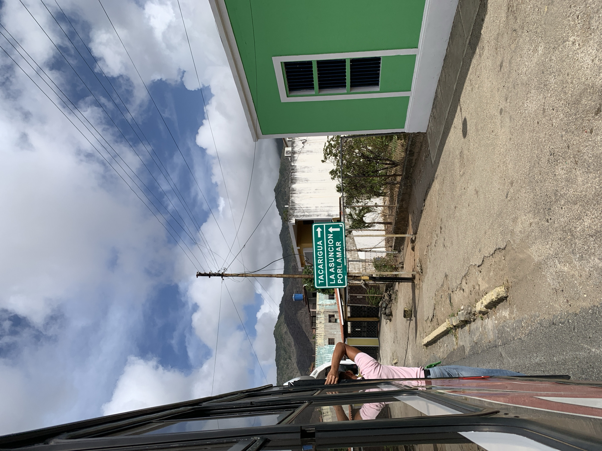 The image is taken from inside a bus, looking out onto a street scene in Tacarigua, Venezuela. A man, wearing a pink shirt and blue jeans, is looking out of the bus window. A green building with a barred window stands adjacent to the cracked concrete road. The street has a signpost pointing in opposite directions for Tacarigua and La Asuncion/Porlamar. Overhead, power lines stretch across a sky with scattered white clouds. A mountain range is visible in the background.