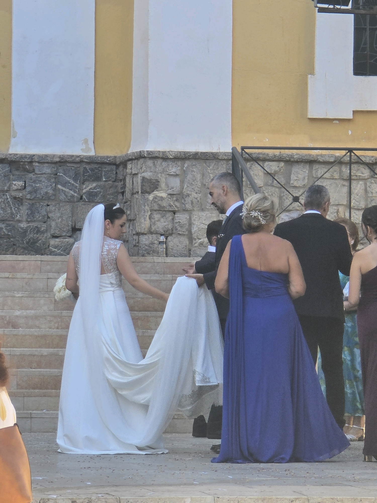In Málaga, Spain, a bride in a white wedding dress with a veil is seen standing on a stone staircase. She is holding a bouquet of white flowers. To her left, a man in a suit is holding the train of her dress. To her right, a woman in a blue one-shoulder dress is standing. In the background, a yellow building with white columns and a stone wall are visible.