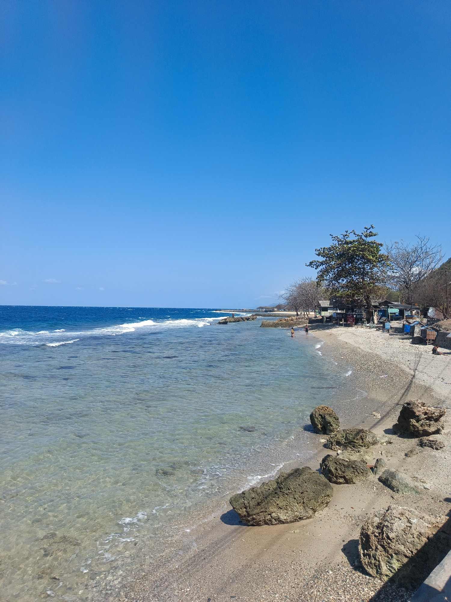 A tranquil, clear-watered beach scene unfolds under a bright blue sky in Botubarani, Indonesia. The pristine water gently laps against a shore composed of fine, light-colored sand and scattered pebbles. Large, weathered rocks are strategically placed along the shoreline, some partially submerged in the shallow water. In the distance, the ocean stretches out, its surface marked by soft white waves.
The beach is sparsely populated. A few individuals are visible near the water's edge, seemingly engaged in casual activities. Further back, a cluster of small, rustic structures are erected, hinting at a local establishment or temporary shelters. A large tree with lush green foliage stands prominently amongst these structures, providing a focal point against the backdrop of a distant, arid hillside. The scene exudes a sense of calm and natural beauty, characteristic of a tropical coastal environment.
