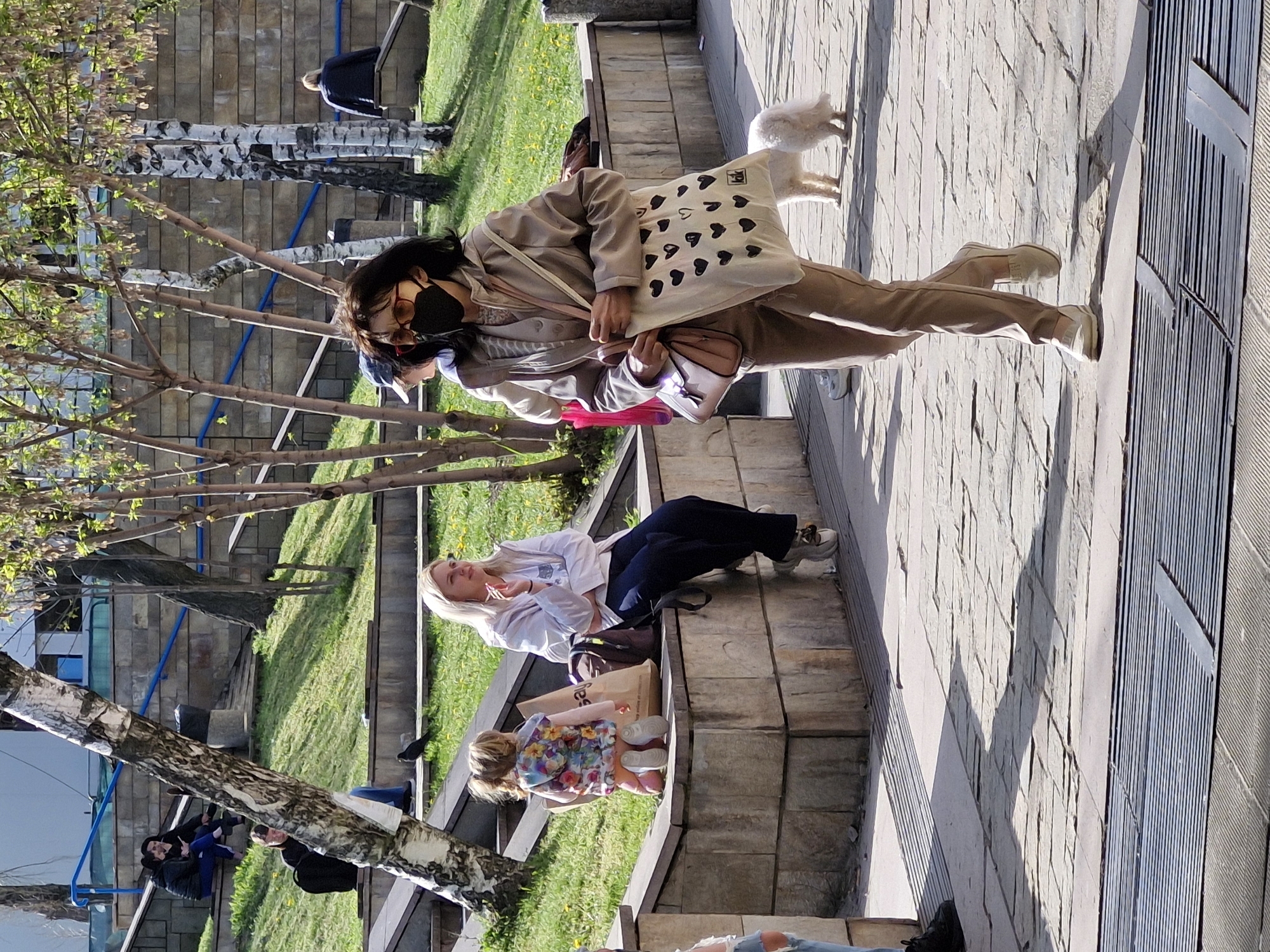 This outdoor image captures a sunny day in what appears to be an urban park or pedestrian area in Sofia, Bulgaria, characterized by paved walkways, green grass, and birch trees with fresh spring foliage. The strong shadows indicate it is daytime, likely mid-morning or afternoon.
In the foreground, a woman with dark hair walks towards the right. She wears a beige jacket, matching trousers, and light-colored flat shoes. Her face is obscured by a black face mask and sunglasses with red frames. She carries a white tote bag adorned with numerous black heart shapes and what appears to be the letters "RK" or "RЯ" in the top right corner, along with a light pink purse slung across her body. A small, fluffy white dog walks closely beside her.
To the left, a blonde woman is seated on a stone bench or raised planter. She wears a white shirt, dark blue pants, and light-colored sneakers, looking upwards and holding a small object. Next to her, a young child in a colorful floral top and light shoes is also seated on the planter, with a brown paper bag visible nearby.
In the mid-ground, other individuals are present: a person in a dark jacket is partially visible behind the blonde woman, and another individual in a dark blue jacket is seen walking further back on the left. In the far background on the right, a person in a black jacket sits on a bench, facing away from the camera. The setting includes architectural elements like stone walls, steps, and visible blue pipes or railings in the background.