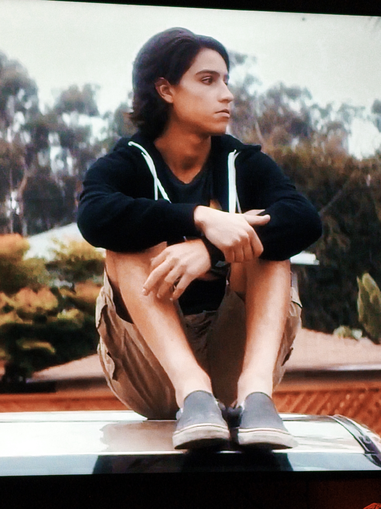 A young male with dark, medium-length hair and brown eyes is sitting on the flat surface of a car. He is wearing a black hoodie with white drawstrings, khaki shorts, and grey slip-on shoes. His arms are crossed, and he is looking to his right, with his head tilted slightly. The background consists of blurred trees and foliage, suggesting an outdoor setting. The overall lighting is soft, and the image appears to be a still from a video or film. The location is not identifiable as Vladivostok, Russia.