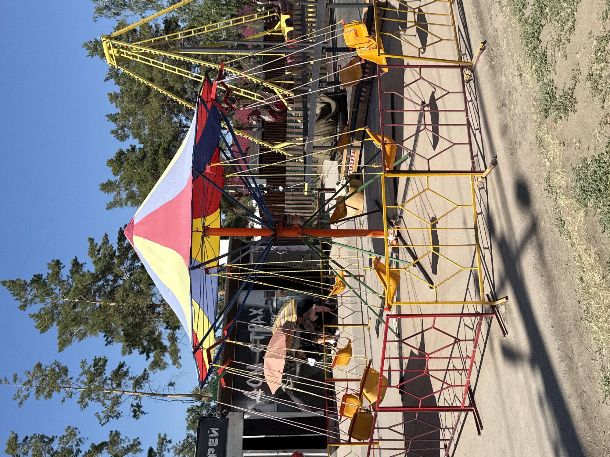 The image depicts a children's swing ride in an amusement park, likely located in Yarovoye, Russia, given the context of the query. The ride features a colorful, multi-sectioned canopy in shades of red, yellow, blue, and white. Suspended from the canopy are several yellow, bucket-style swings with chains. The ride is enclosed by a fence made of yellow and red metal bars with a diamond pattern. In the background, there are tall pine trees under a clear blue sky. Behind the swing ride, a larger, yellow amusement ride structure with a ladder-like appearance is visible. To the left of the swing ride, there is a building with dark walls and some graffiti, featuring Cyrillic text. Underneath the swing ride's canopy, two individuals are seated, partially obscured by an umbrella, suggesting a pause or rest. The ground is a mixture of paved and dirt areas with sparse vegetation. The scene is illuminated by sunlight, casting distinct shadows.