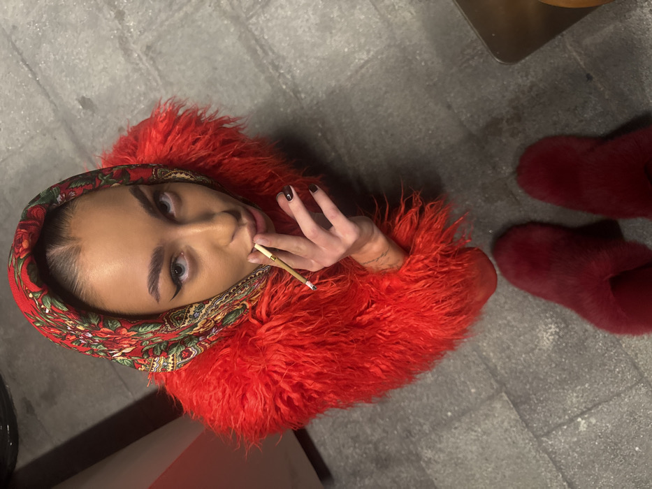 A female individual is depicted from a high-angle perspective, looking directly upward into the camera. She wears a red, green, and gold patterned headscarf and a voluminous red shag coat. Her eyes are light-colored, and she has dark winged eyeliner. Her right hand, featuring dark-painted fingernails and forearm tattoos, holds a lit, hand-rolled cigarette with a filter tip positioned near her mouth. Below her, a pair of red fuzzy slippers are visible. The subject stands on a paved surface composed of dark grey rectangular tiles. A partial, dark, possibly metallic object is present on the ground to the far right. This scene is located in the city of Dubai, United Arab Emirates.