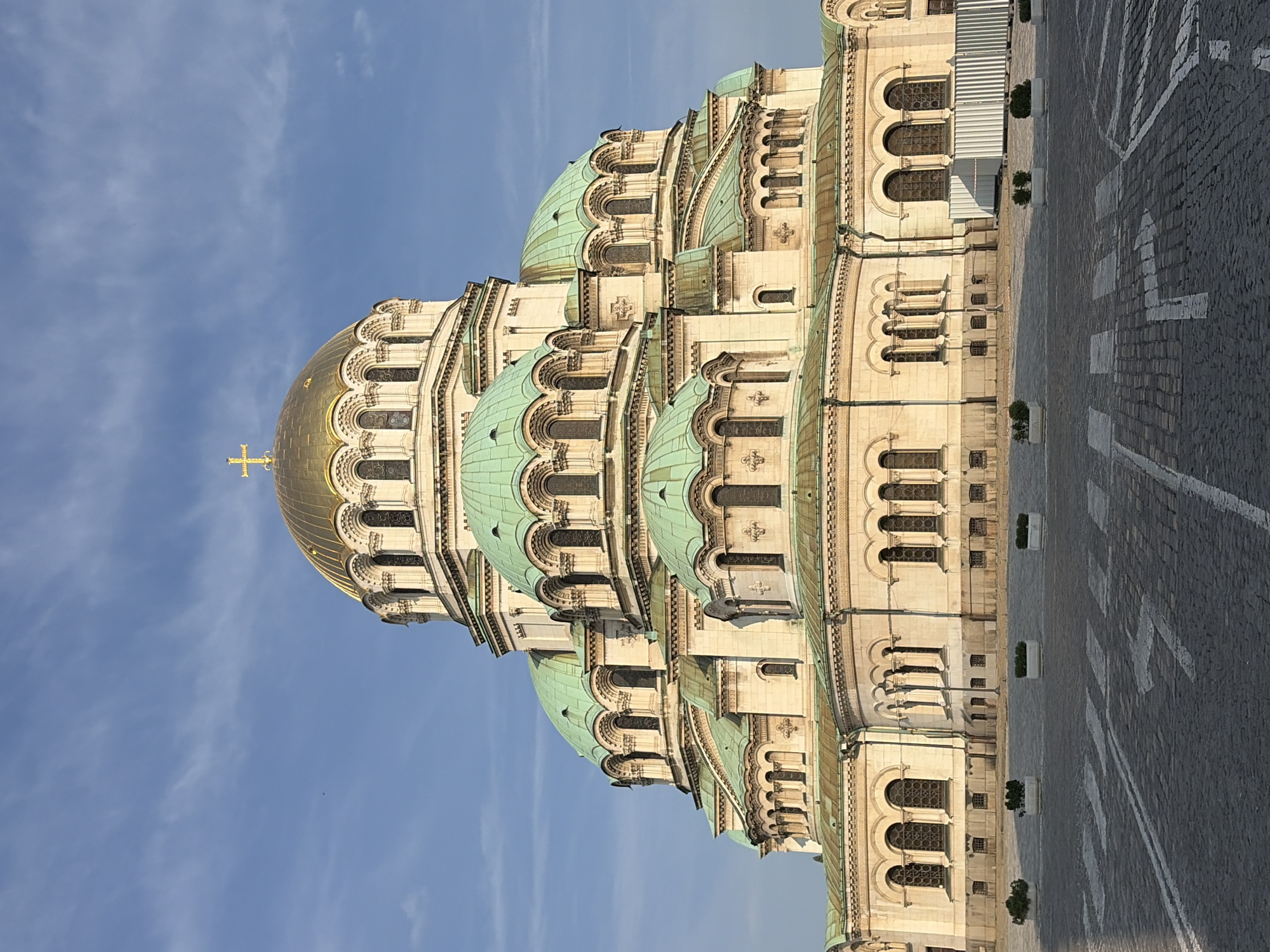 The image displays a detailed frontal view of the Alexander Nevsky Cathedral, located in Sofia, Bulgaria. The central architectural feature is a large golden dome surmounted by a golden cross, with numerous smaller green-patinated copper domes arranged in tiered layers around the main structure. The cathedral's facade is composed of light-colored stone or stucco, featuring arched windows, decorative cornices, and architectural detailing.
The foreground consists of a cobblestone plaza marked with white crosswalk stripes and white directional arrows. Four white rectangular planters, each containing small green foliage, are positioned in a linear arrangement on the plaza. No individuals are present. On the right side of the image, a dark street lamp is partially visible, positioned near the cathedral's base, and a section of a white corrugated metal barrier is also partially observable. The sky above is clear blue with sparse, high-altitude white clouds. The scene is illuminated by natural light, casting subtle shadows on the building and ground.
