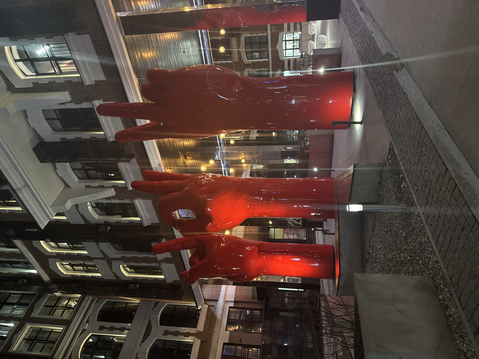 An exterior nighttime image depicts three oversized, glossy red hand sculptures illuminated by red uplights in an urban courtyard in Moscow, Russia, specifically within the Hampton by Hilton Moscow Strogino complex or the adjacent "Rechniki" residential development. From left to right, the sculptures display a "rock and roll" gesture, an "OK" gesture, and a "peace" gesture. These installations are positioned on a paved area consisting of cobblestones and smooth, light-colored pavers, flanked by concrete planters containing sparse, bare vegetation. In the background, modern multi-story buildings with dark brick facades, large windows, and white architectural trim are visible, with some interior lights active. Reflective architectural elements are also present. No individuals are observed. Ground-level bollard lights provide supplementary illumination to the scene.