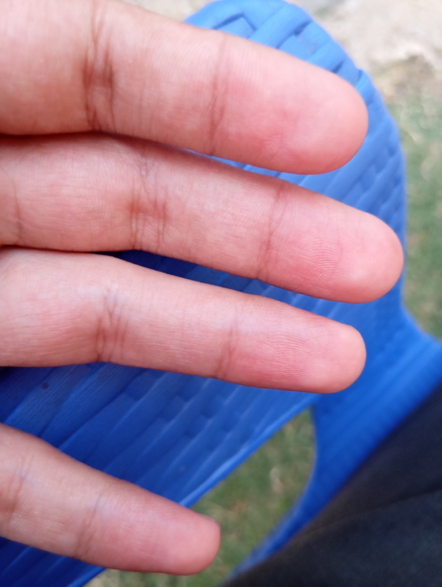 The image is a close-up shot of a person's hand, with several fingers visible. The hand is resting on a bright blue, woven-textured object, which appears to be a chair or seating surface. The fingers are slightly curved and show visible skin texture, including faint lines and creases. The background is blurred, with hints of green grass and possibly some light brown earth or foliage, suggesting an outdoor setting. A dark fabric, likely clothing, is visible in the lower right corner of the frame. The lighting appears natural, casting soft shadows and highlighting the skin tones of the hand. There is no visible text in the image. The overall impression is that of a casual, relaxed moment, possibly outdoors on a sunny day.