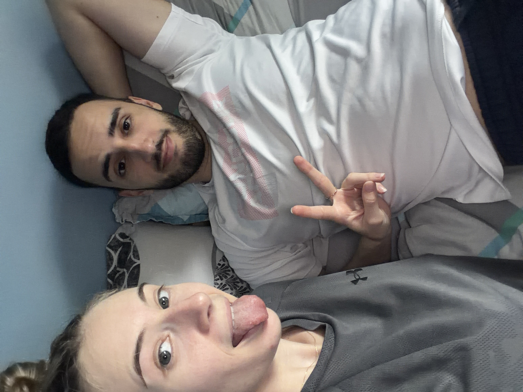 Two individuals are lying on a bed. One individual is male, with dark hair and a beard, wearing a white t-shirt and black pants. The other individual, presumably female, has blonde hair and is wearing a grey athletic shirt and is sticking her tongue out. The male is holding up two fingers in a peace sign. The setting appears to be indoors, on a bed with a patterned pillow and blanket. There is no indication of the location in Plovdiv, Bulgaria.