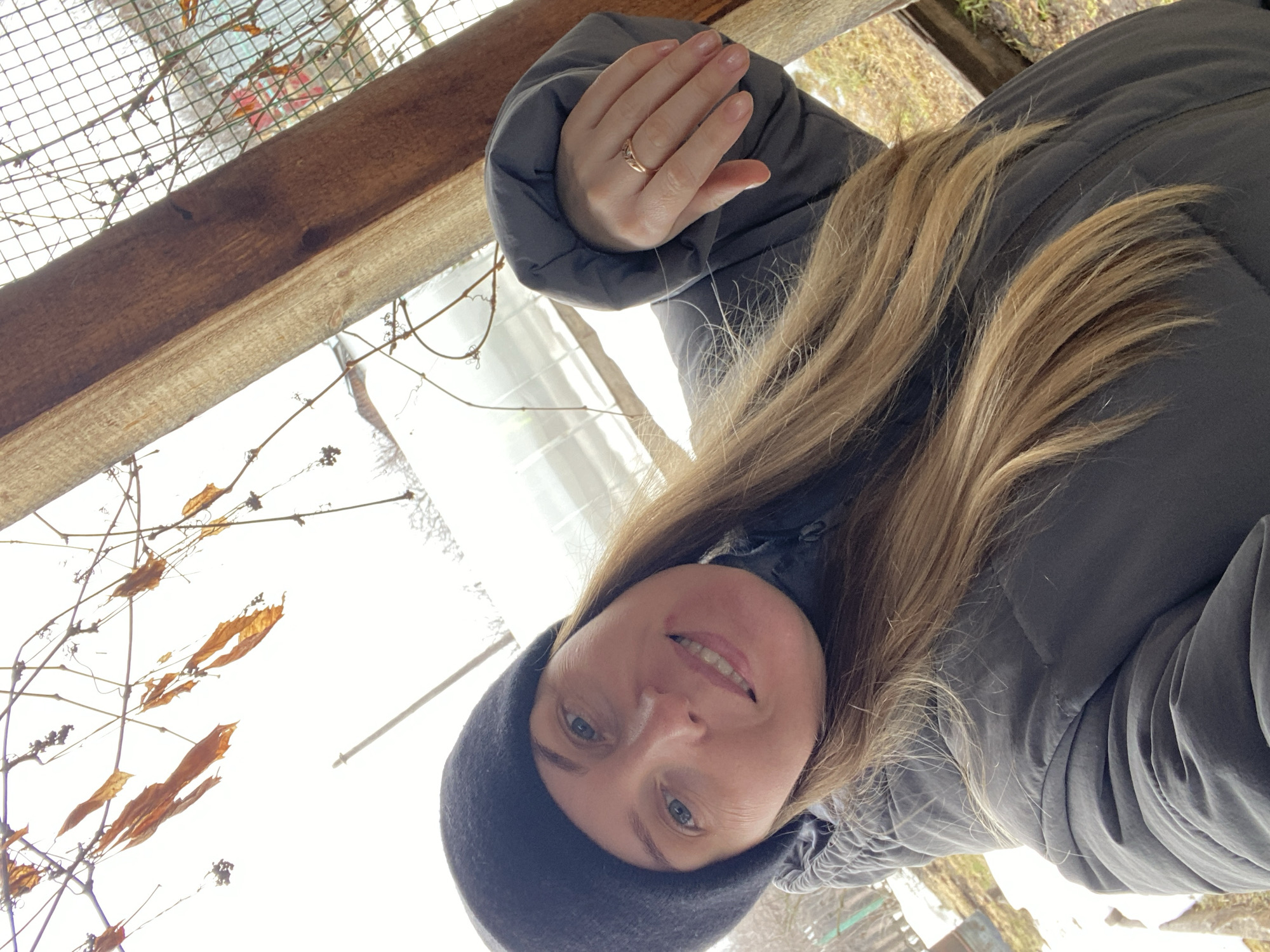 A woman with blonde hair, wearing a black beanie and a gray puffer jacket, is taking a selfie. She has a gold ring on her left ring finger. The background appears to be outdoors, with a wooden structure, bare branches, and some foliage. The location is likely Chaikovsky, Russia, based on the implied context, but that is not definitively visible in the image itself. The woman is smiling.