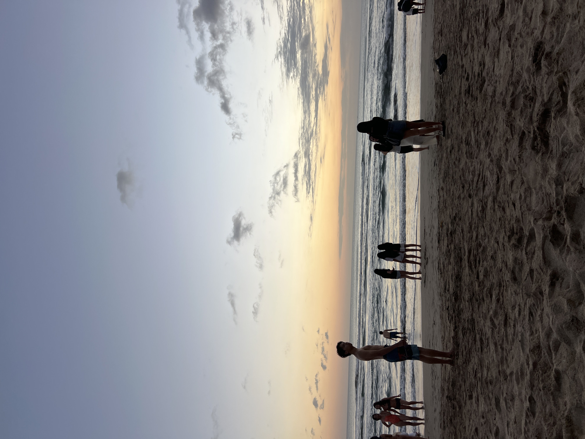A serene beach scene at dusk in Kuta, Indonesia, features a group of individuals on the sand and in the shallow water. The sky transitions from a pale blue at the top to a warm gradient of yellow and orange near the horizon, with scattered clouds. Waves roll gently onto the shore. In the foreground, dark sand is visible with footprints. Several people are standing on the wet sand near the water's edge. A young man, shirtless and wearing patterned swim trunks, stands in the foreground, facing to his left. Further back, a group of women and a man are present in the water. A woman in a patterned swimsuit and another in dark attire are near the water. To the right, a group of women stand together, one of whom appears to be taking a photo. A dark bag or shoe rests on the sand in the mid-ground. The overall atmosphere is calm and suggestive of the end of a day at the beach.