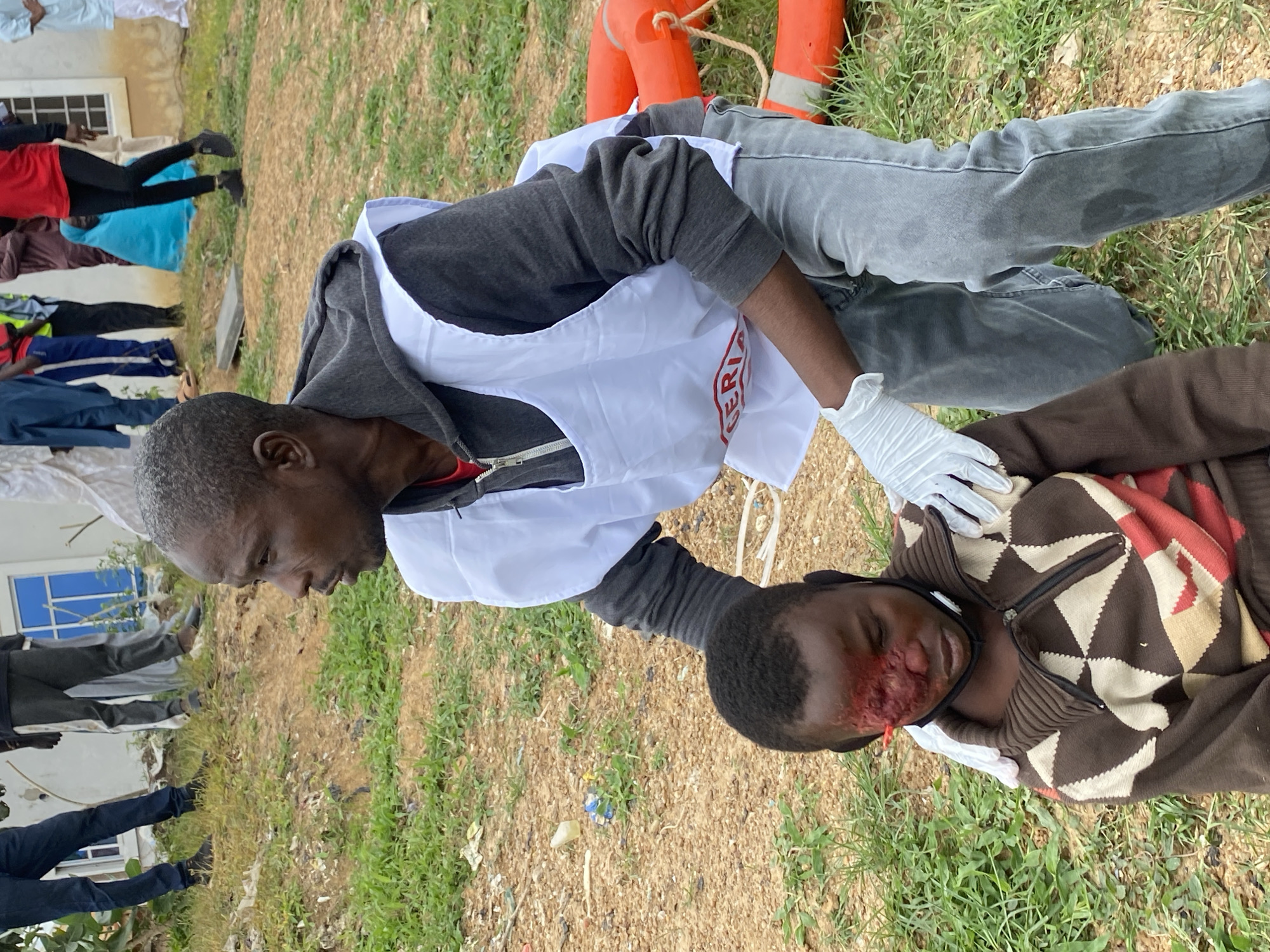 In Kawo, Nigeria, a man wearing a white vest with red lettering and grey pants crouches down to attend to a young boy who is seated. The boy has a severe facial injury on the left side of his face, near his eye and nose, which is red and appears to be bleeding. The man is wearing white gloves and is reaching towards the boy's head. The environment is outdoors with dry, reddish-brown earth, sparse green grass, and some debris scattered on the ground. In the background, other individuals are visible, some standing, and a building with blue-paned windows can be seen. Two orange life rings are also present on the ground to the right of the main subjects.