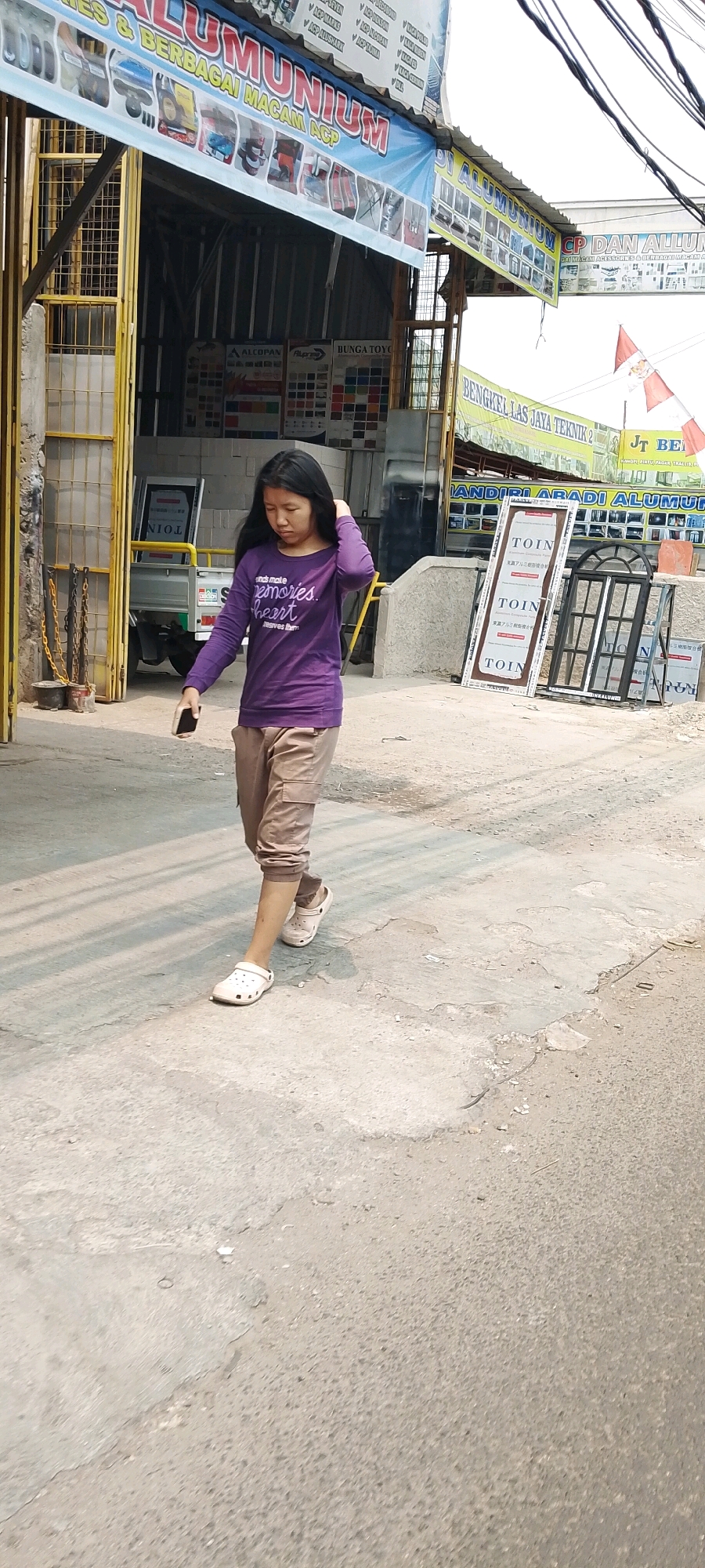 A young woman with long, dark hair walks along a paved street in front of a store in Bekasi, Indonesia. She wears a purple long-sleeved shirt with white lettering that reads, "friends make memories, heart leaves them." Her pants are beige cargo pants, and she wears white slip-on shoes. The woman holds a dark-colored smartphone in her right hand, and her left hand is on her head, touching her hair. The store behind her appears to sell aluminum products and features several signs with Indonesian text, as well as window frames and displays. The sun casts shadows on the street, indicating it is daytime.