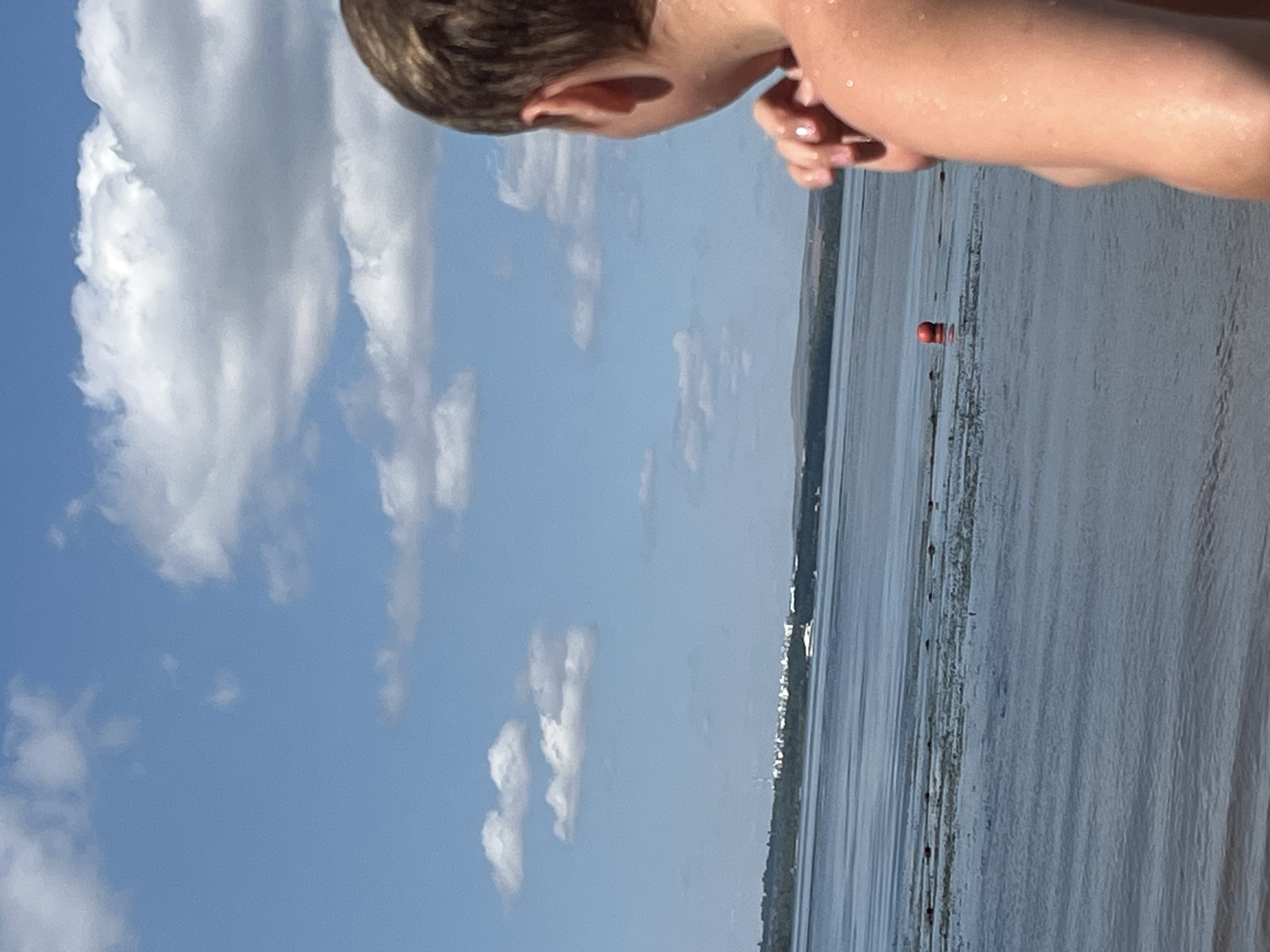 The image shows a close-up view of the back of a young person's head and shoulder, looking out at a body of water. The person appears to be on a beach or at the edge of a lake. The water is calm and reflects the blue sky, which is dotted with white, fluffy clouds. In the distance, across the water, there is a shoreline with trees and a cluster of white buildings. A small white boat is visible on the water, and a red buoy floats near the shore. The lighting suggests it is a sunny day. The person's skin is wet, indicating they may have been swimming. The image is shot from a low angle, looking up towards the sky. No specific landmarks in the city of Shumeika, Russia are identifiable.