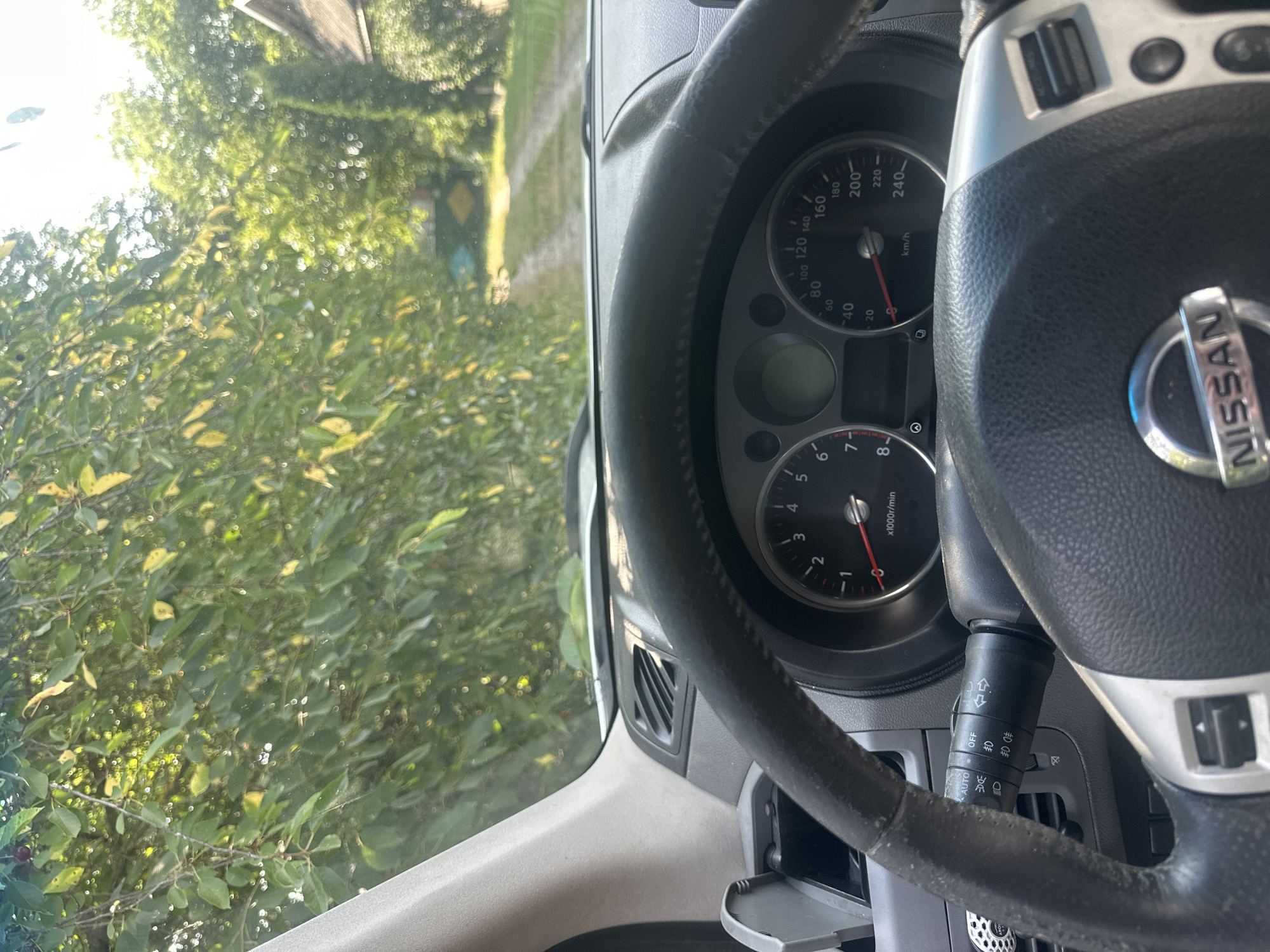 This is a first-person view from inside a Nissan vehicle, parked on a dirt road lined with lush green trees and foliage. The dashboard, including the steering wheel and instrument cluster, is visible. The speedometer indicates a speed of approximately 0 km/h, and the tachometer shows around 800 rpm, suggesting the engine is idling. The car appears to be stationary on an unpaved road, possibly in a rural area. The background shows a dirt path leading into a densely vegetated area with trees and a small, partially visible building to the right. The presence of trees and the nature of the road suggest a location outside of a dense urban environment, potentially in or near Shatava, Ukraine, given the context provided. There are no people visible in the image. The scene depicts a car parked in a natural, outdoor setting.