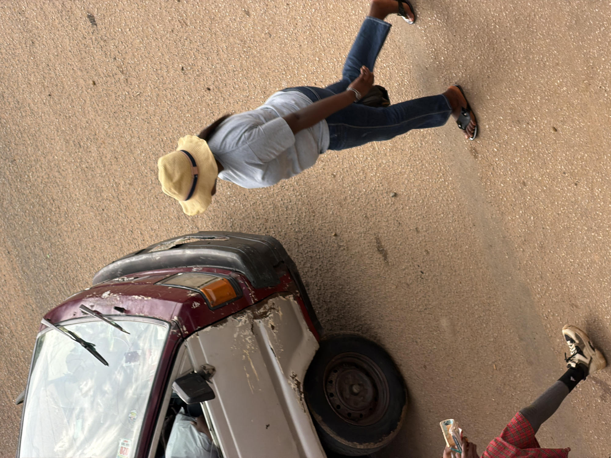 The image depicts an outdoor scene with a vehicle and two individuals. A maroon and white vehicle, appearing to be a van or small bus with significant body damage and rust, is parked at an angle. A person wearing a yellow straw hat, a light blue t-shirt, and dark blue jeans is walking away from the vehicle. They are wearing dark sandals. To the left of the vehicle, another person, partially visible, is wearing a red and black checkered skirt, a dark grey leg covering, a black sock, and a white and black athletic shoe. This individual is holding what appears to be a small packet or box. The ground is a reddish-brown, sandy or gravelly surface, suggesting an unpaved area or road. The scene is likely a roadside or informal gathering space in Bukuru, Nigeria.