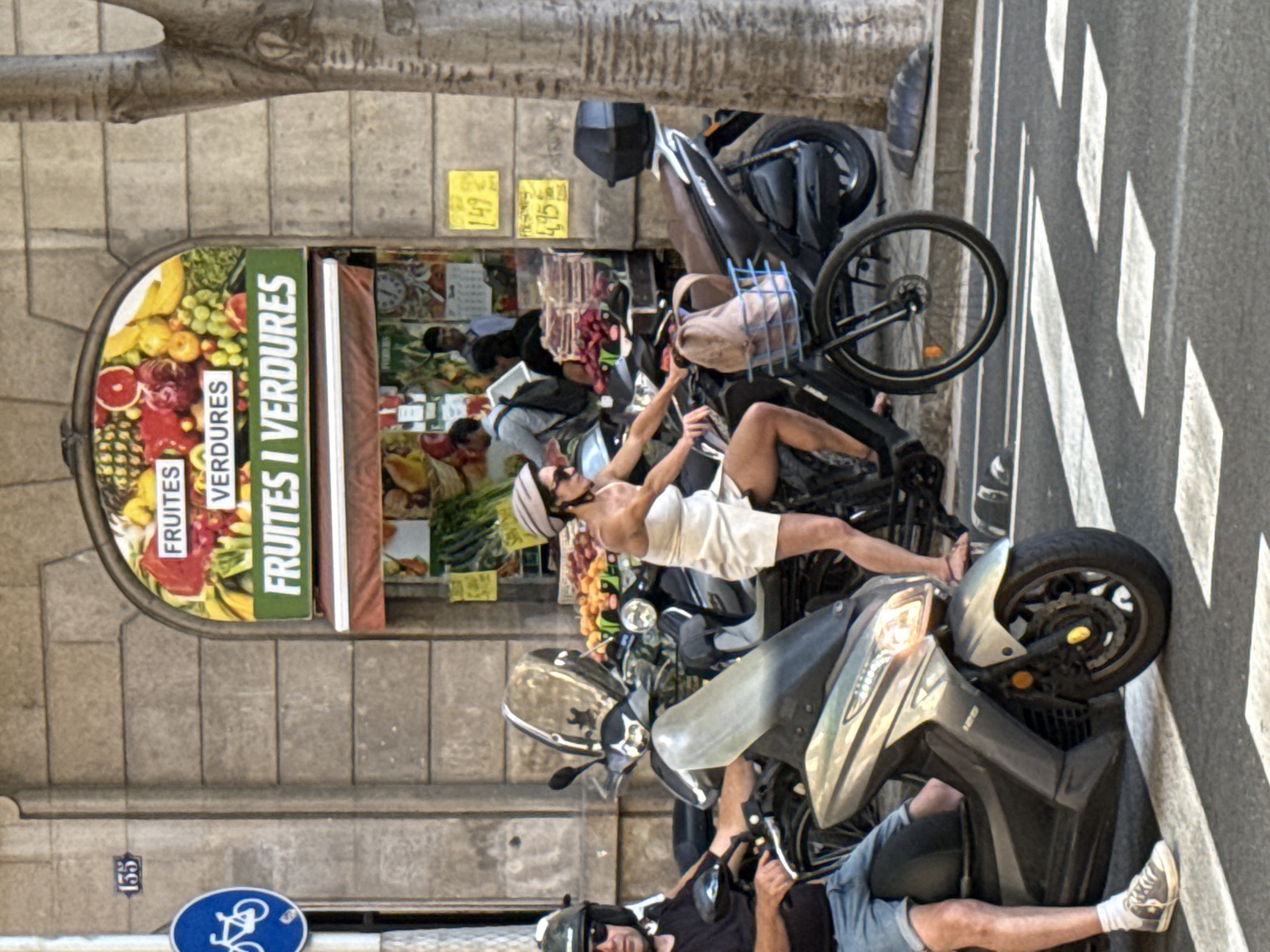 The image depicts a street scene in Barcelona, Spain, likely on a sunny day. In the foreground, two individuals are on scooters, stopped at a crosswalk. The person on the left, a male wearing a black t-shirt, blue shorts, and a helmet, is positioned on a silver scooter. The person on the right, a female wearing a light-colored off-the-shoulder dress and a helmet, is on a black scooter. She is also on a bicycle that is integrated with the scooter in some way.
Behind the scooters, a fruit and vegetable store is visible with a prominent arched sign that reads "FRUITES I VERDURES" (Fruits and Vegetables in Catalan). The store's facade is decorated with images of various fruits. The street is paved, with white markings indicating a crosswalk. A tree trunk is visible on the right side of the image, and a building with stone cladding forms the background. There are price tags visible on the building's exterior, with prices like "1.49" and "4.95". A blue circular sign with a white bicycle symbol is mounted on the building to the left.