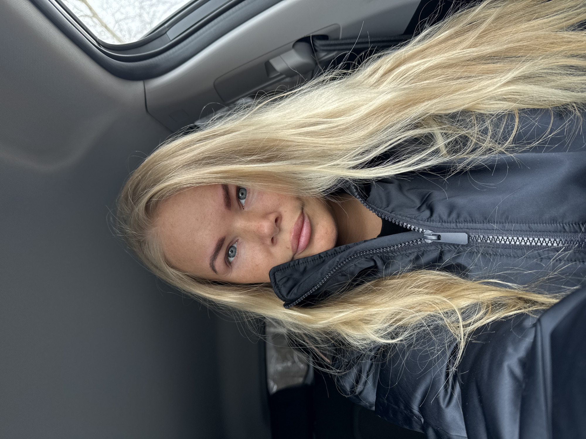 A blonde woman with light blue eyes is looking at the camera. She is wearing a black puffy coat. She is sitting in a car. The car has a sunroof. The city is Unknown, Unknown.