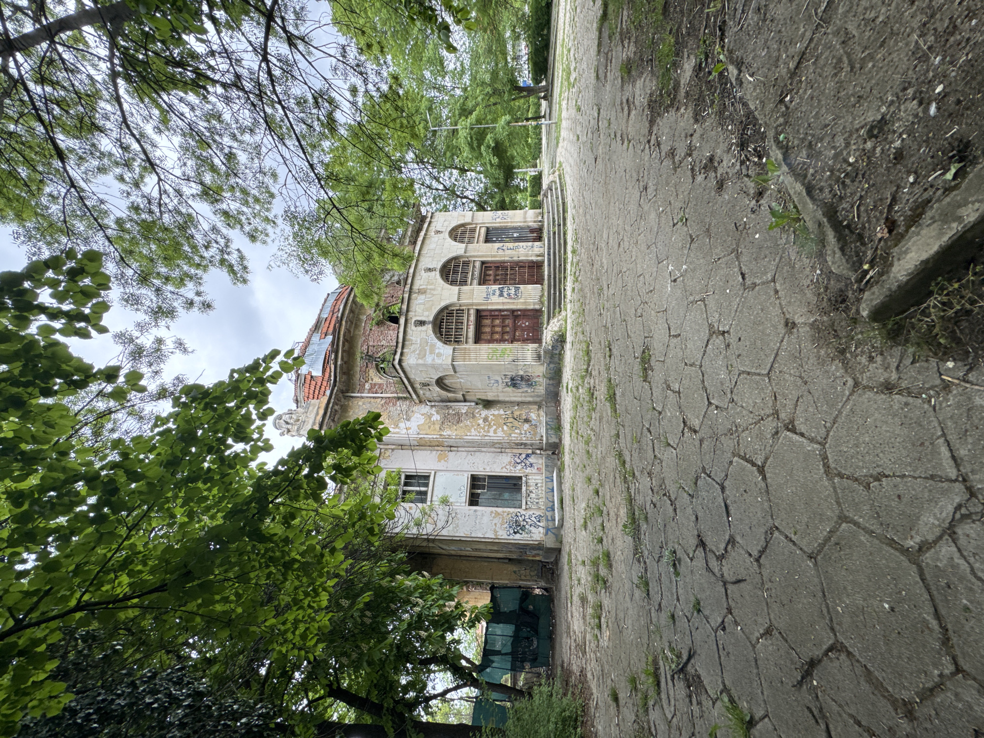 An eye-level, angled outdoor photograph depicts an aged building from a side perspective in Sofia, Bulgaria. The building appears abandoned, with a crumbling facade, and is covered in graffiti. The roof is partially damaged. The entrance features three arched doorways with barred windows. A set of concrete steps leads to the entrance.
To the right of the building is a paved walkway with geometric concrete blocks, suggesting a public park or garden setting. Weeds are growing in the cracks of the pavement.
Trees with full green leaves obscure parts of the building and surround the area. The sky is overcast.
The photograph's angle creates a sense of perspective, with the walkway and building receding into the distance. There are no people visible in the shot.