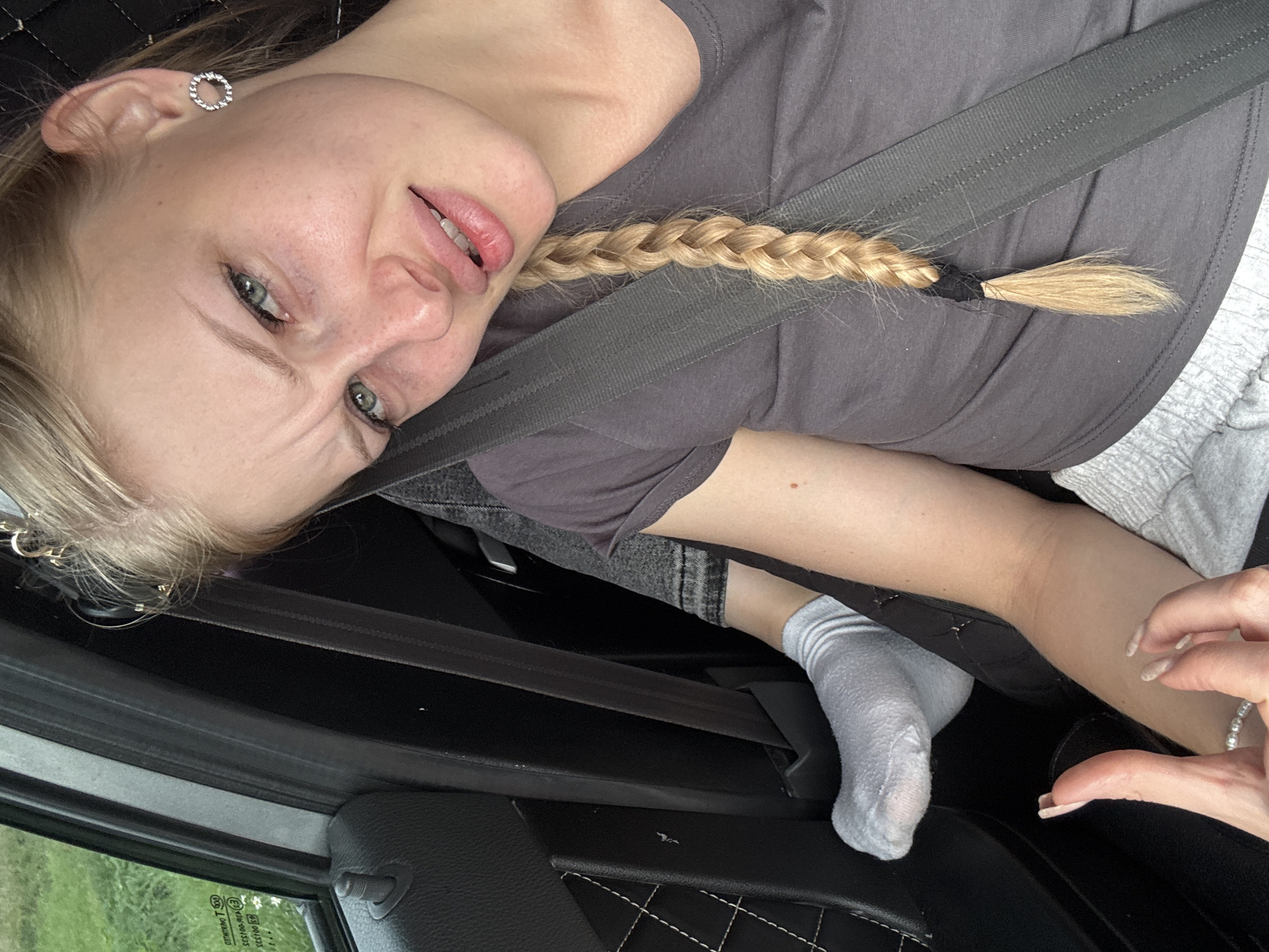 A young woman with blonde hair braided down her back is sitting in the passenger seat of a car. She is wearing a grey t-shirt and grey sweatpants. A seatbelt is fastened across her chest. Her left foot is visible, wearing a white sock. She has a facial expression of discomfort or skepticism. The background shows the interior of a car, including the door panel and window, with greenery visible outside. The location is not specified in the image or provided text.