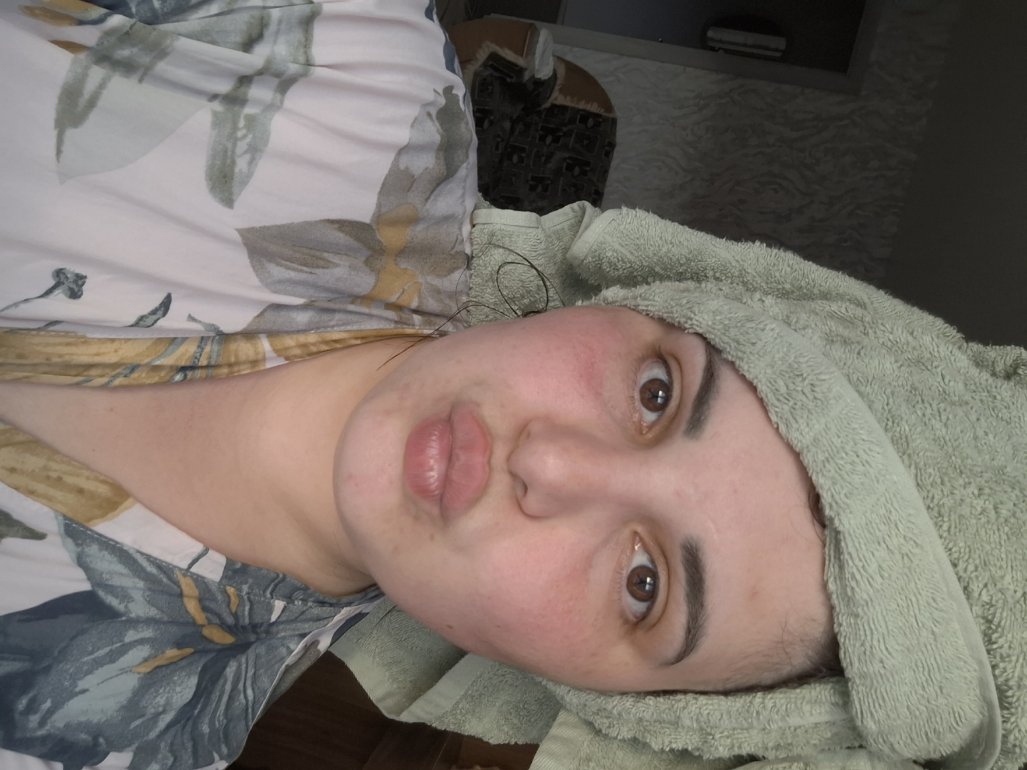 A person is lying down, with a green towel wrapped around their head. They have fair skin, brown eyes, and are making a kissing face. The person is wearing a floral print top. The location appears to be indoors, with a patterned wall and furniture visible in the background. There is no indication this is in the city of Амзя, Russia.
