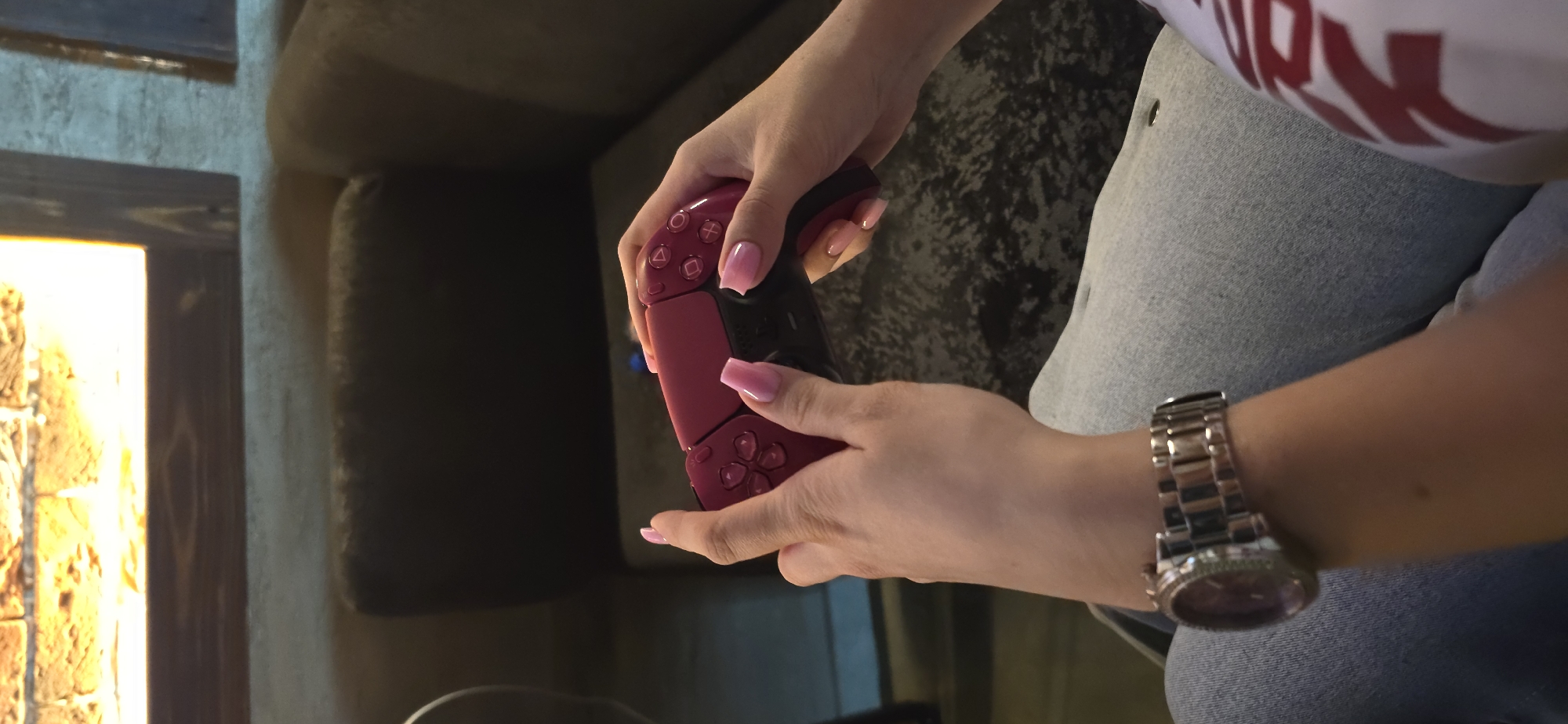 The image shows a close-up of a person holding a red video game controller. The person's hands have manicured nails with a pink gradient design. They are wearing a silver watch with a large face on their left wrist and a grey denim garment. In the background, a dark couch is visible, and to the left, a fireplace with glowing embers and a wooden frame is partially visible. The lighting suggests it might be evening or indoors, creating a cozy atmosphere. The specific location is Krasnodar, Russia.
