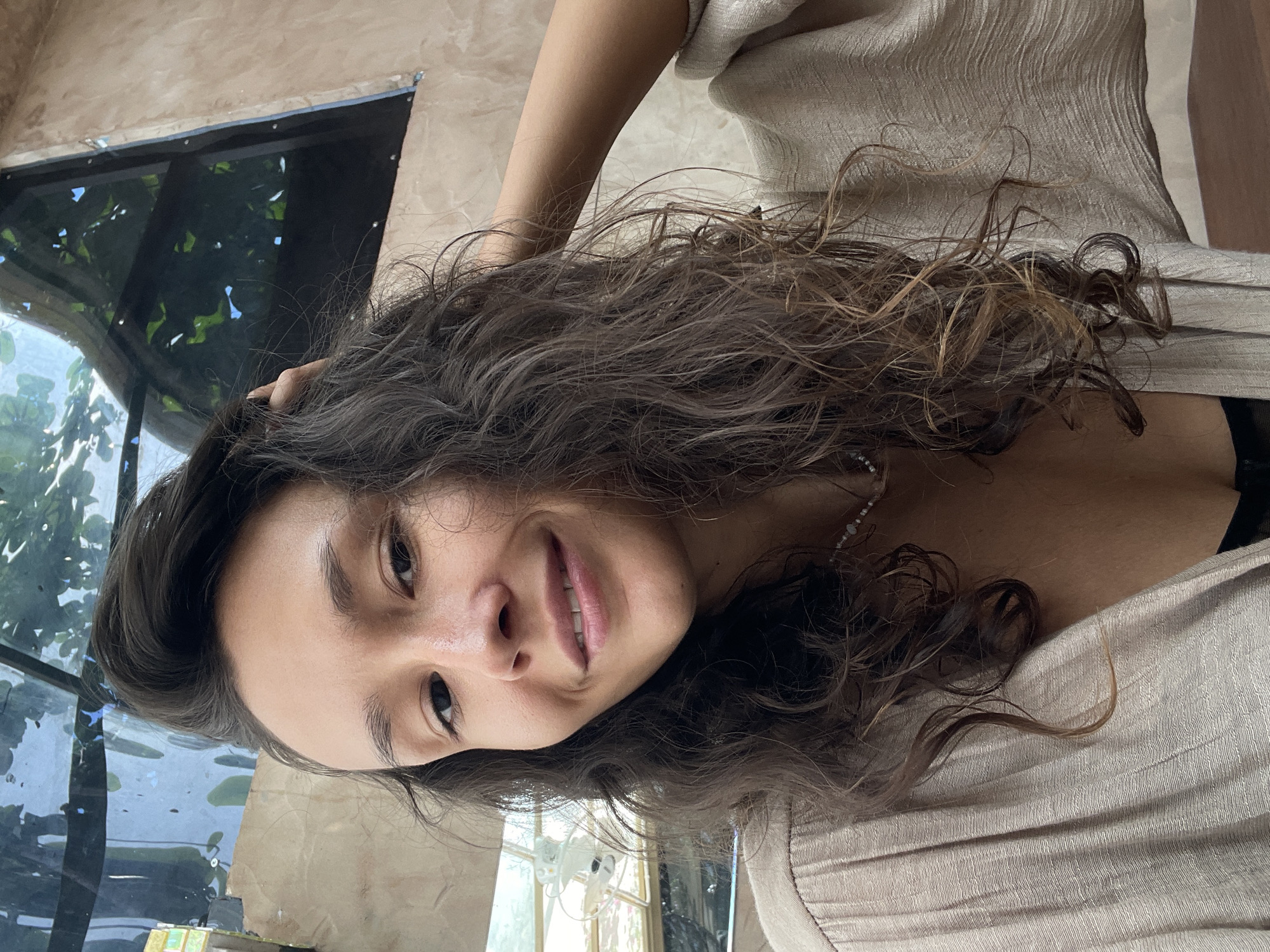 A young woman with long, dark, wavy hair, featuring some lighter brown highlights, smiles gently at the camera. Her head is tilted, and her right hand is lightly placed in her hair near her head. She wears a light-colored, possibly beige or cream, textured top and a delicate necklace. The setting appears to be an indoor or semi-enclosed space, with large windows or glass panels in the background. Through these windows, lush green foliage is visible, indicating an outdoor view of a tropical environment, consistent with the Mũi Né, Vietnam location context. The interior wall behind her is light brown and subtly textured. The natural, bright lighting suggests it is daytime. The woman's expression conveys calmness and friendliness. The original image is rotated 90 degrees counter-clockwise. There is no visible text.