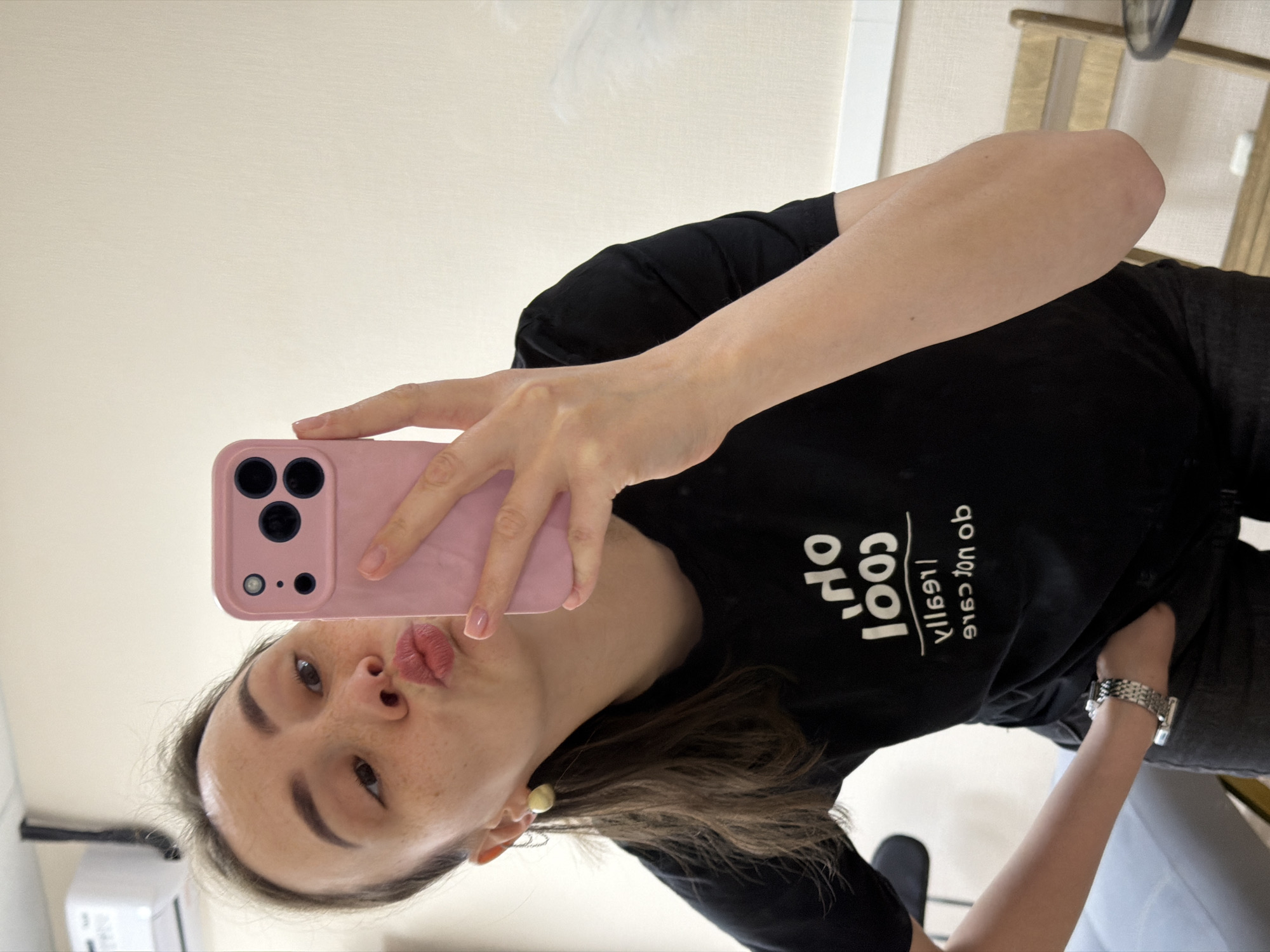 A person takes a selfie with a pink phone case. The person has brown hair, freckles, and wears a black t-shirt with white text. They have light skin, full lips, and an earring. The location appears to be indoors with beige walls and furniture. The city is not discernible from the image.