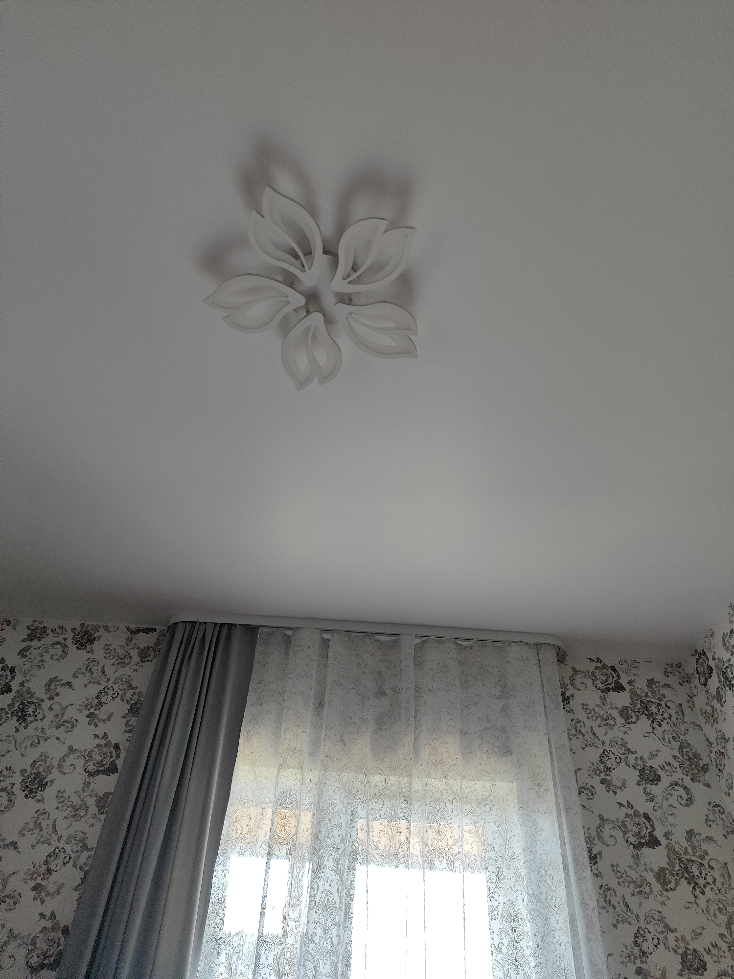 A ceiling fixture shaped like a flower is mounted on a white ceiling. Below the ceiling, a window is covered by a sheer, patterned curtain and a thicker, solid grey curtain. The walls are wallpapered with a floral design. The scene appears to be an interior of a room.