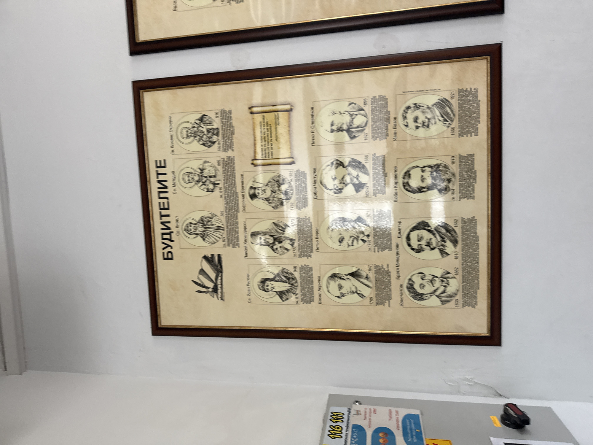 A framed, sepia-toned poster titled "БУДИТЕЛИТЕ" (Awakeners/Educators) hangs on a white wall in what appears to be an indoor public or educational setting in Varna, Bulgaria. The poster features a grid of portraits of significant Bulgarian historical figures, including saints, writers, and educators, arranged chronologically.
The figures depicted include Saints Cyril, Methodius, Clement of Ohrid, and John of Rila, representing early spiritual and literary awakeners. Later figures such as Paisiy Hilendarski, Sophronius of Vratsa, Vasil Aprilov, Petar Beron, Dobri Chintulov, Petko R. Slaveykov, the Miladinov Brothers (Konstantin and Dimitar), Lyuben Karavelov, and Ivan Vazov are also featured, with their birth and death years indicated.
In the center of the poster, a scroll graphic contains a quote in Bulgarian, partially visible, which states: "Sacred is the deed of those who lit the fire of national education and carried the national spirit through the dark centuries of slavery."
Below the main poster, another white board or display is visible, with text including "116 111" and "Национална телефонна линия..." (National telephone line...), suggesting a helpline, possibly for children ("Училищна" - School). The overall display serves as a tribute to Bulgarian cultural and national heroes.