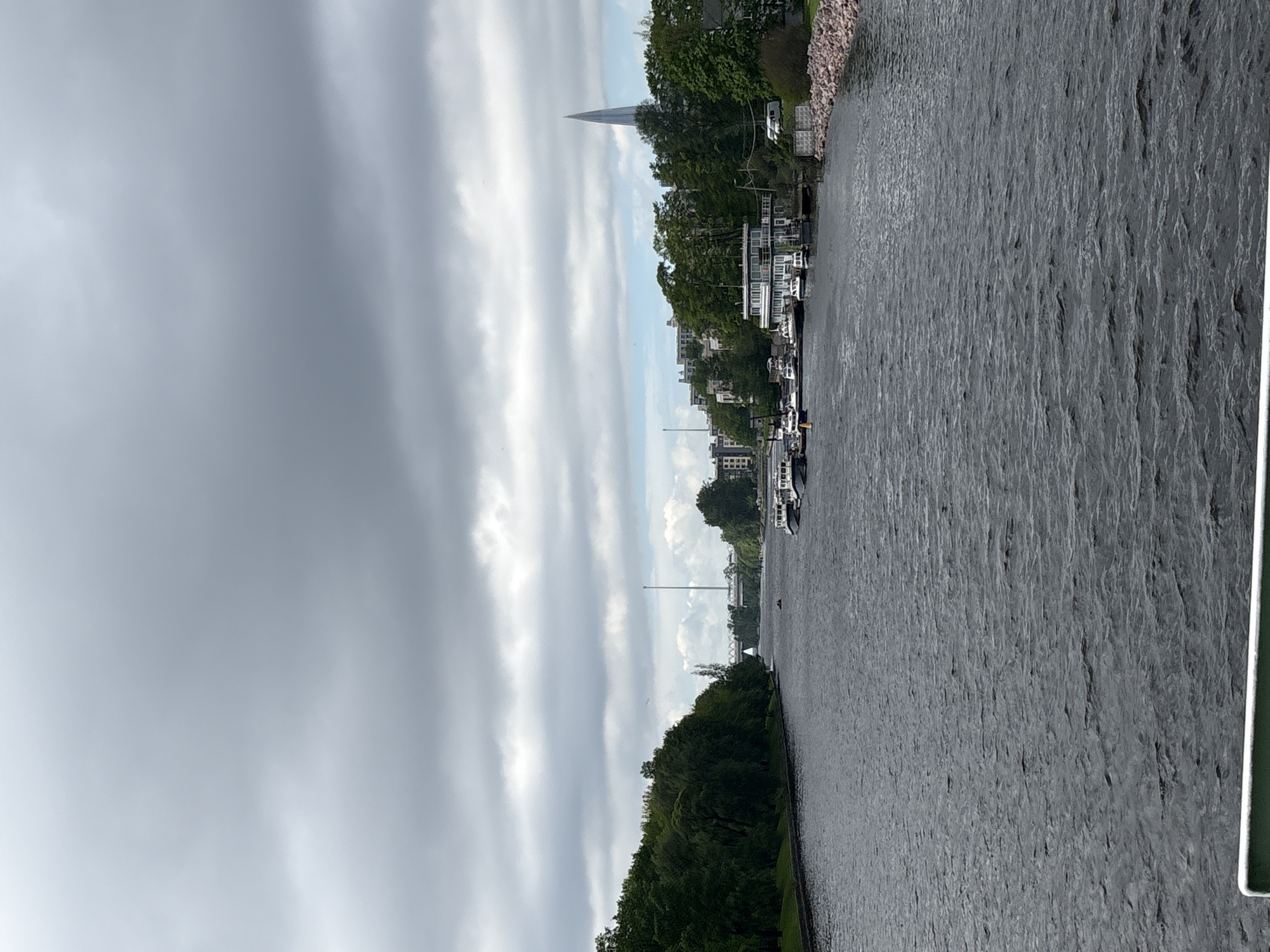 The image depicts a waterscape along a canal or riverbank in Saint Petersburg, Russia, characterized by a dense overcast sky. The water surface shows visible ripples and texture, reflecting the ambient light.
Along the right side, a stretch of vegetation lines the riverbank, transitioning into residential buildings with docks and boats moored alongside. A slender spire or tower is visible in the distance.
The overall mood is subdued due to the heavy cloud cover, giving the scene a somewhat gray and muted appearance. The frame includes part of a structure, possibly a bridge or embankment, in the lower right corner.