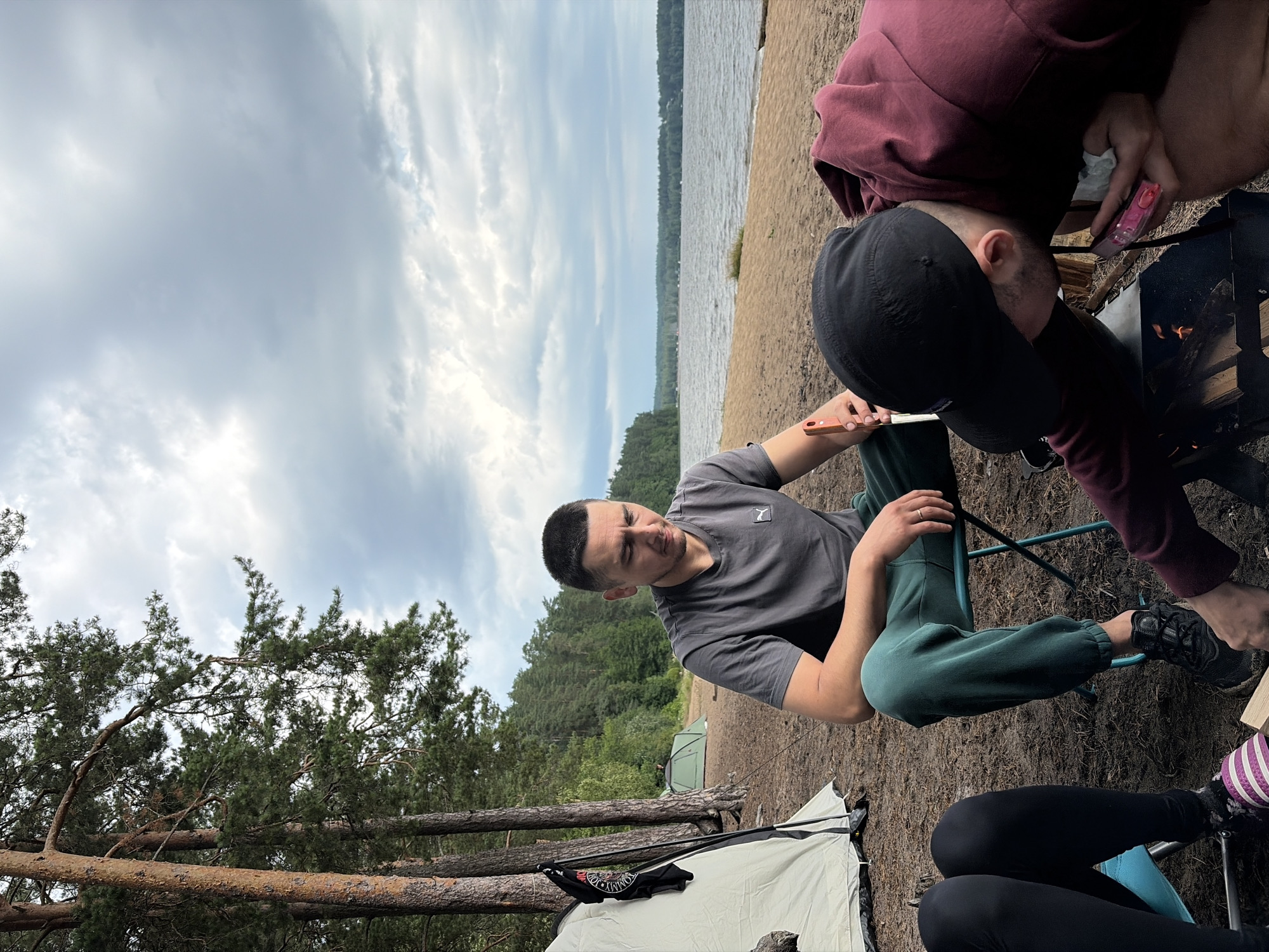 The image depicts a group of people engaged in a camping activity near a lake in Vaskelovo, Russia. In the foreground, a man sits on a blue folding chair, holding a knife. To his right, another person, wearing a black cap and a maroon hoodie, is leaning over a campfire. A tent is visible to the left, and another green tent is set up in the background. The environment features a sandy beach, a lake, and a forested area with pine trees. The sky is overcast with dramatic clouds.
