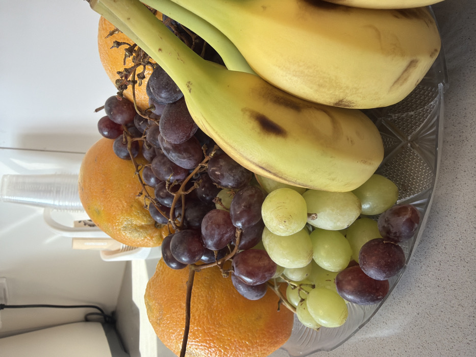 The image displays a clear glass fruit bowl filled with various fruits, situated on a light grey, speckled countertop in a domestic kitchen setting within the Bnei Shimon Regional Council, Israel. The bowl, which has a textured pattern and scalloped rim, contains several oranges, bunches of dark red/purple grapes, clusters of light green grapes, and at least three yellow bananas with minor brown markings. In the background, a white wall is visible, along with part of a white kitchen appliance with a grey base on the far left. Towards the center-back, behind the fruit, a white holder containing multiple clear plastic cups and what appears to be brown stirrers or utensils is present. The scene is illuminated by natural light, casting subtle shadows. No individuals are present, and no actions or interactions are depicted beyond the arrangement of the fruit.
