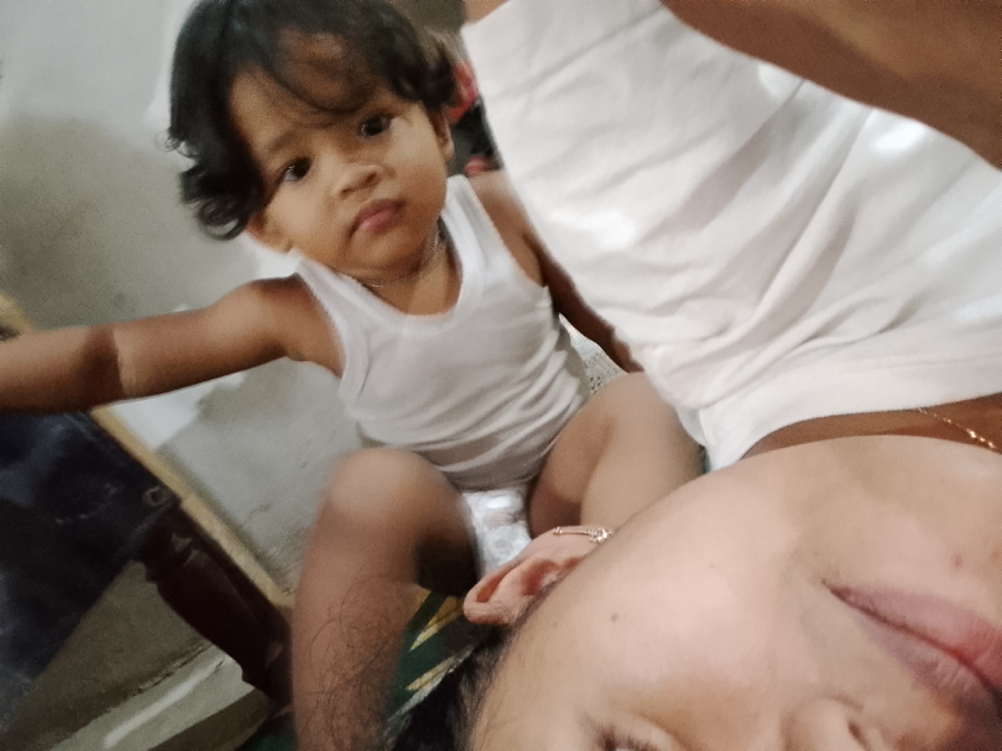 In Besuki, Indonesia, a close-up, low-angle selfie shows a toddler seated and looking towards the camera with a neutral expression. The toddler is wearing a white tank top. In the foreground, the face of the person taking the selfie is visible, tilted downwards. They are wearing a white top, and a thin gold necklace is visible around their neck. Part of a wooden chair or furniture is visible on the left side of the frame. The background is a plain, light-colored wall.