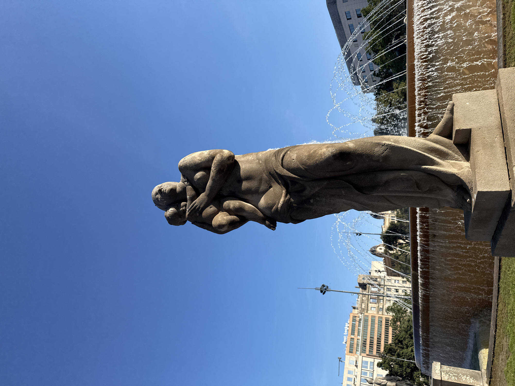 The image shows the Fountain of a Mother, located in Barcelona, Spain. It depicts a stone sculpture of a mother holding a child. The fountain water is visible. Buildings and trees are in the background. The sky is clear and blue.