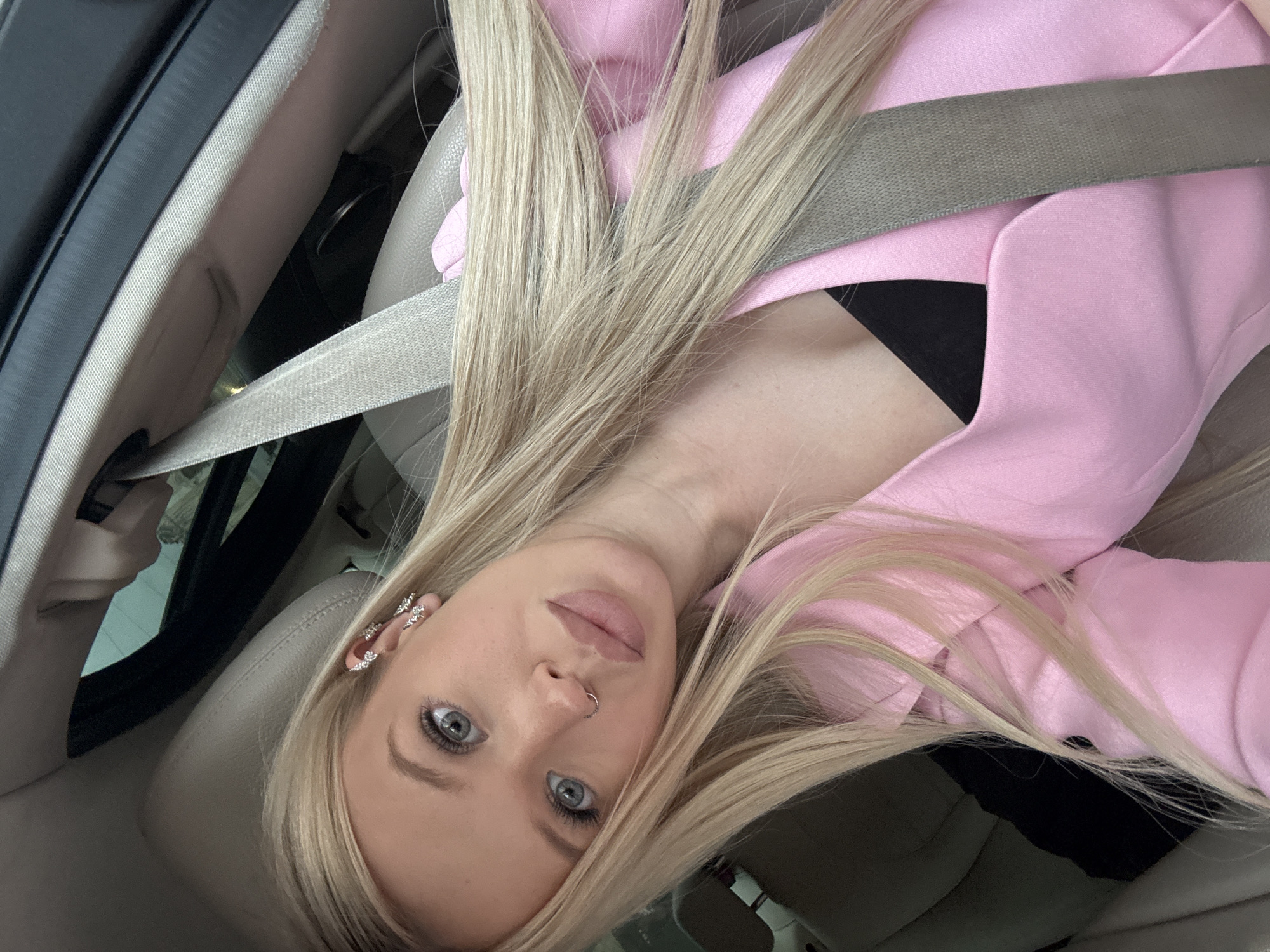 This is a selfie taken inside a car, likely in Saint Petersburg, Russia. The subject is a young woman with long blonde hair, blue eyes, and light skin. She is wearing a pink blazer over a black top and has several pieces of jewelry, including earrings and a nose ring. Her hair is obscuring part of her face and is spread across her shoulder and chest. A seatbelt is clearly visible across her torso, indicating she is the driver or a passenger in the vehicle. The interior of the car is visible, with a light-colored leather seat and part of the car door. The lighting suggests it could be daytime, though the exact time is not discernible. The overall mood appears relaxed and casual. There is no visible text in the image.