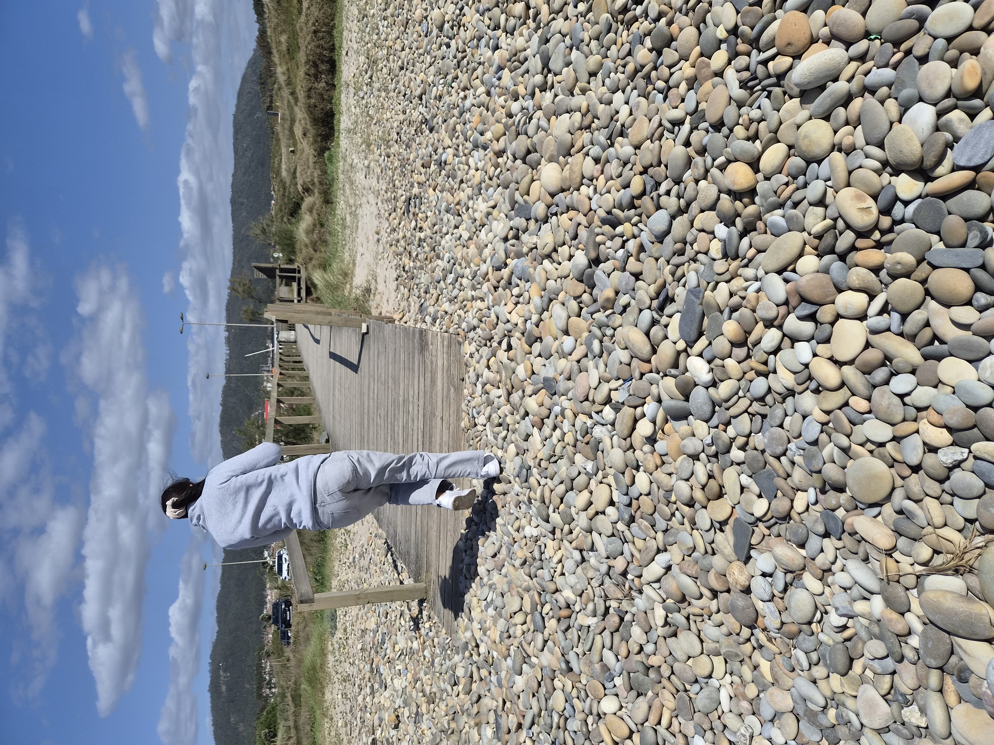 A horizontal, eye-level shot shows a person standing on a wooden walkway along a rocky beach. The person wears a light gray hoodie and matching pants with white sneakers. They face the background, their head turned slightly to the left. The walkway has wooden posts and sits between the rocks and sandy dunes covered in vegetation. In the background, a hillside rises under a clear blue sky with scattered clouds. Streetlights are visible along the pathway, and there are cars parked in the distance. This scene is located in Esposende, Portugal.