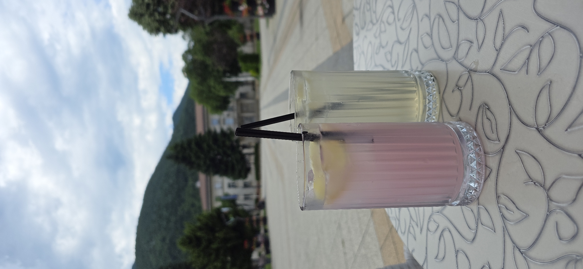 An outdoor, eye-level shot captures two tall, fluted glass tumblers containing cold beverages on a patterned table in the city of Unknown, Unknown. The left glass holds a pink liquid with ice and a lemon slice, while the right contains a pale yellow liquid with ice. Both glasses have a single black straw inserted. The glasses are sharply in focus, exhibiting condensation. The table features a light-colored surface with an intricate, silver-grey embossed or etched floral/leaf pattern.
The background is uniformly blurred, suggesting a shallow depth of field. Behind the drinks, a wide, light-colored paved plaza extends into the distance. Beyond the plaza, a multi-story building with a light facade and a reddish-brown roof is visible, flanked by numerous green trees, including tall conifers. Flags are partially discernible on poles near the building. Further in the background, large, green, tree-covered mountains rise under a partly cloudy sky, with patches of blue visible amidst white and grey clouds. No people are present in the scene. The overall lighting is bright, indicating daytime.