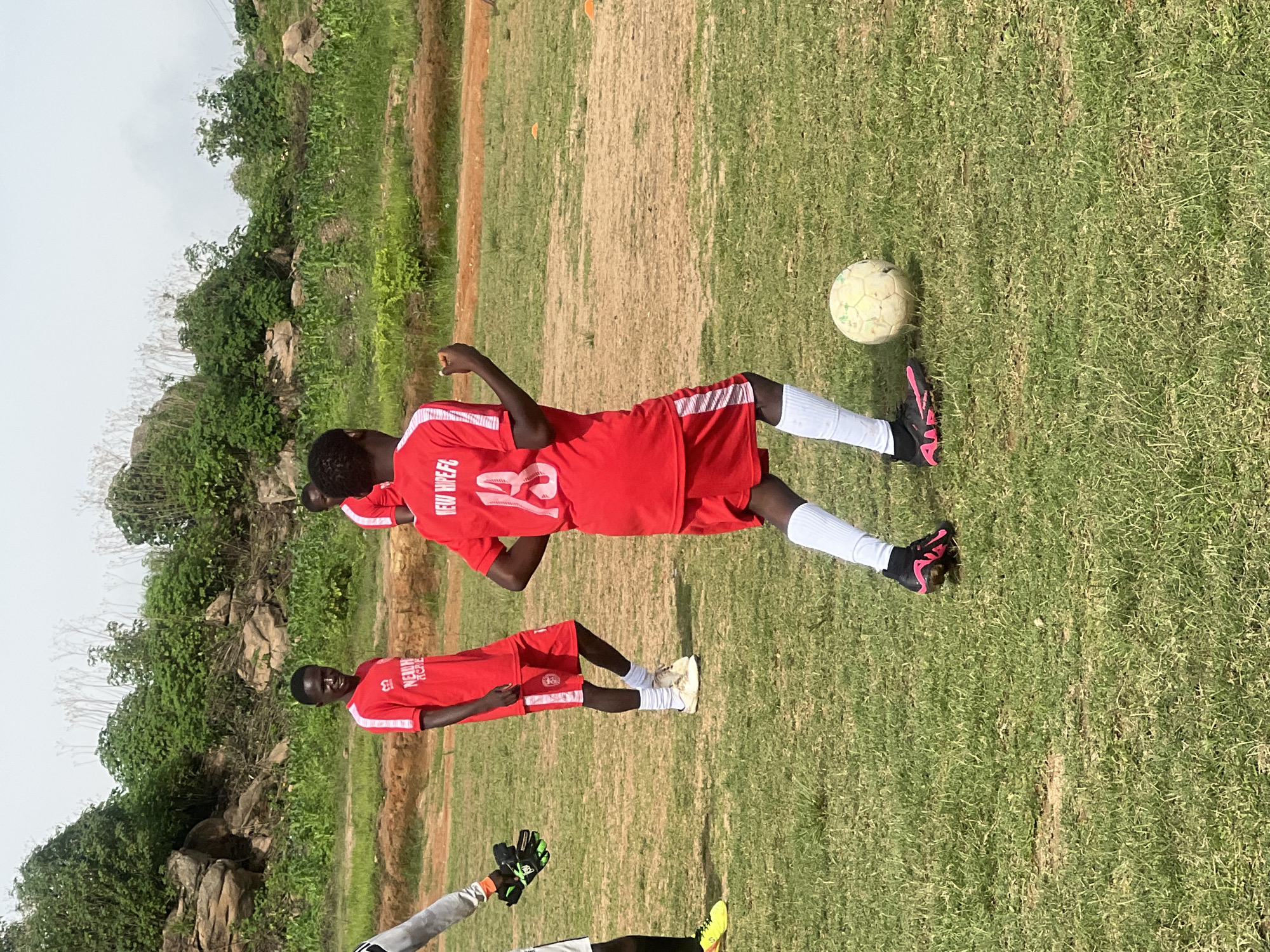 In Jos, Nigeria, on a dry grassy field, three young males in red soccer uniforms are engaged in a soccer practice. The player in the foreground, facing away from the camera, wears a red jersey with the number "13" and the text "NEW HIPE FC". He is dribbling a weathered soccer ball, his right foot poised to kick it. Another player, to the left and further back, also in a red uniform, watches. To the far left, a fourth individual, wearing a grey long-sleeved shirt and black goalkeeper gloves, is visible from the chest down, positioned as if to defend a goal. In the background, a rocky hillside covered in green vegetation and sparse trees forms the backdrop under a hazy sky.