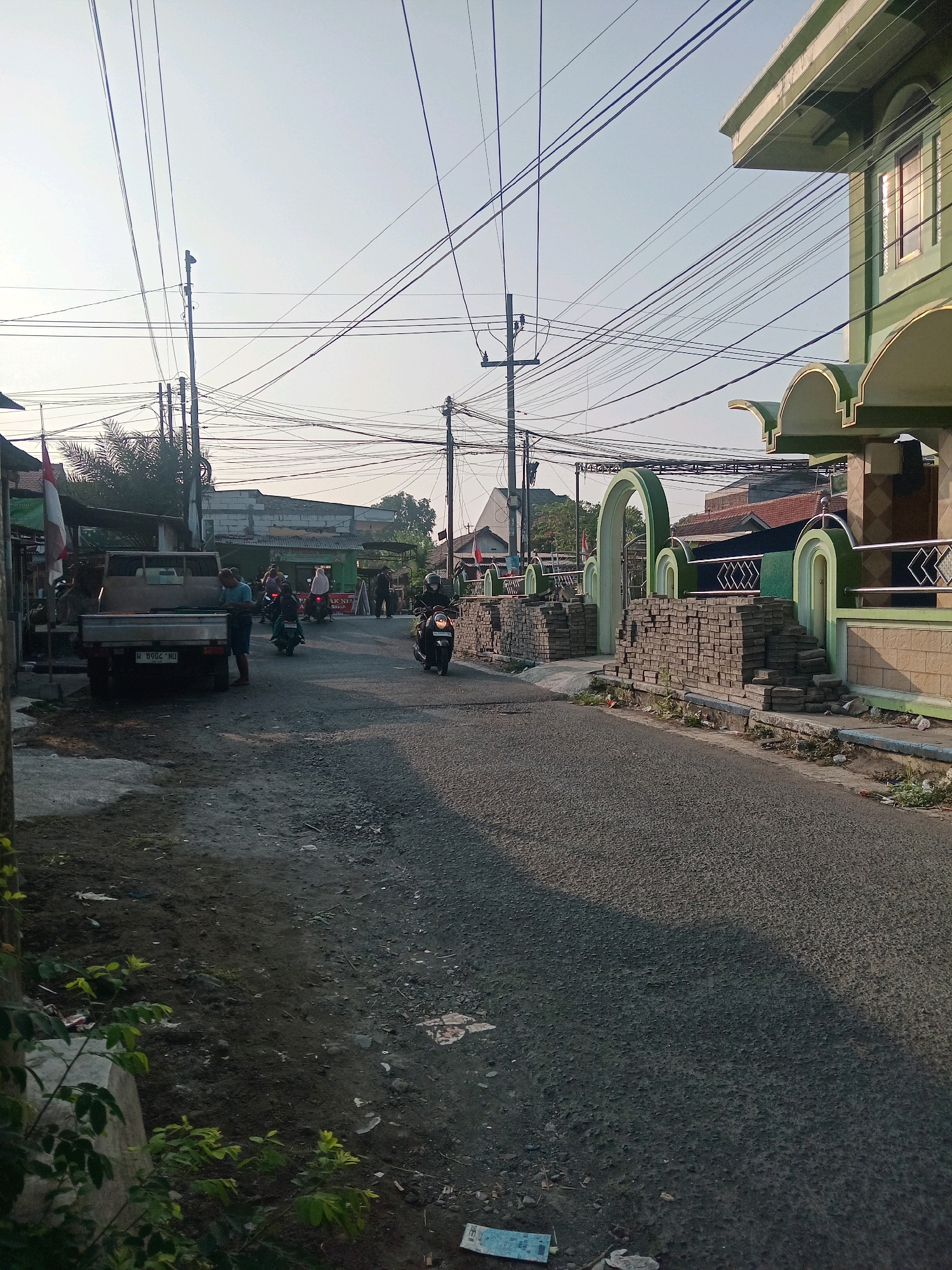 The image depicts a street scene in Banjarsari, Indonesia, with a clear, overcast sky. The road is paved with asphalt and gravel, with a dirt shoulder on the left side. A light truck is parked on the left, facing away from the camera, with its bed loaded with what appears to be building materials. Two men are standing near the truck, one looking towards the truck and the other towards the road. Further down the street, several motorcycles are visible, with riders on some of them. A building with a green facade and arched entrances is situated on the right side of the road, with stacks of grey paving stones in front of it, suggesting ongoing construction or renovation. Overhead, a complex network of electrical wires is strung between utility poles. In the background, there are more buildings and vegetation, consistent with a residential or mixed-use urban environment. An Indonesian flag is visible on a pole on the left side of the image. The overall impression is of a typical, moderately busy street in a tropical urban setting, with elements of daily life and construction activity.