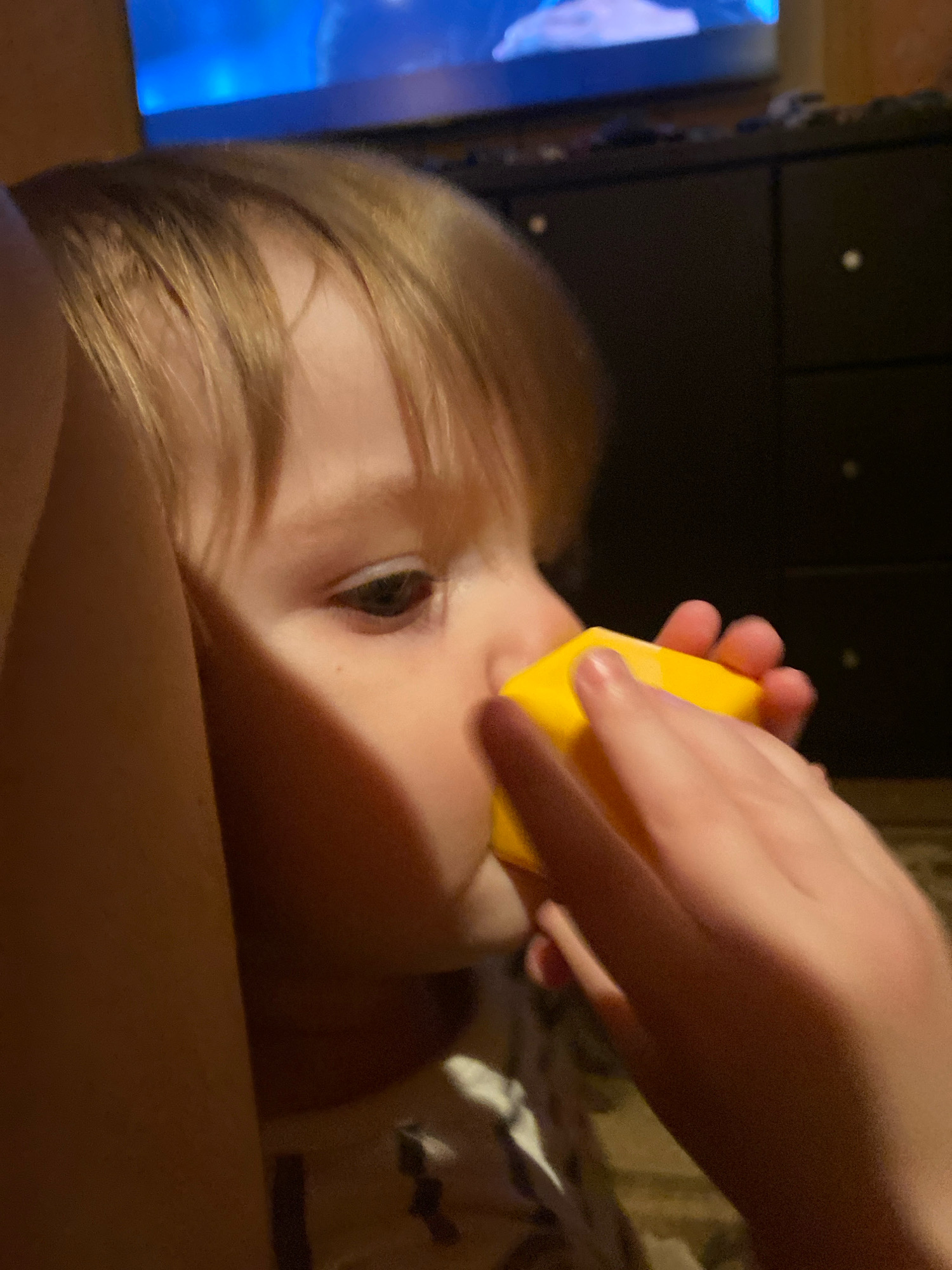 A close-up shot shows a young child, whose hair is light brown and appears to be cut short. The child is looking to the right, with their eye visible. A bright yellow object, possibly a toy block, is held up to their face, with their nose and mouth in close proximity to it. Two hands are visible, one on each side of the yellow object, appearing to be holding it or guiding it towards the child's face. The background is dark and out of focus, showing hints of what might be furniture, including what looks like a dresser with drawers, and possibly a television screen in the upper left corner displaying a blue light. The lighting appears to be dim, suggesting an indoor setting, possibly in the evening or a room with artificial lighting. The overall mood conveyed by the image is one of focused curiosity or play. No visible text is present in the image.