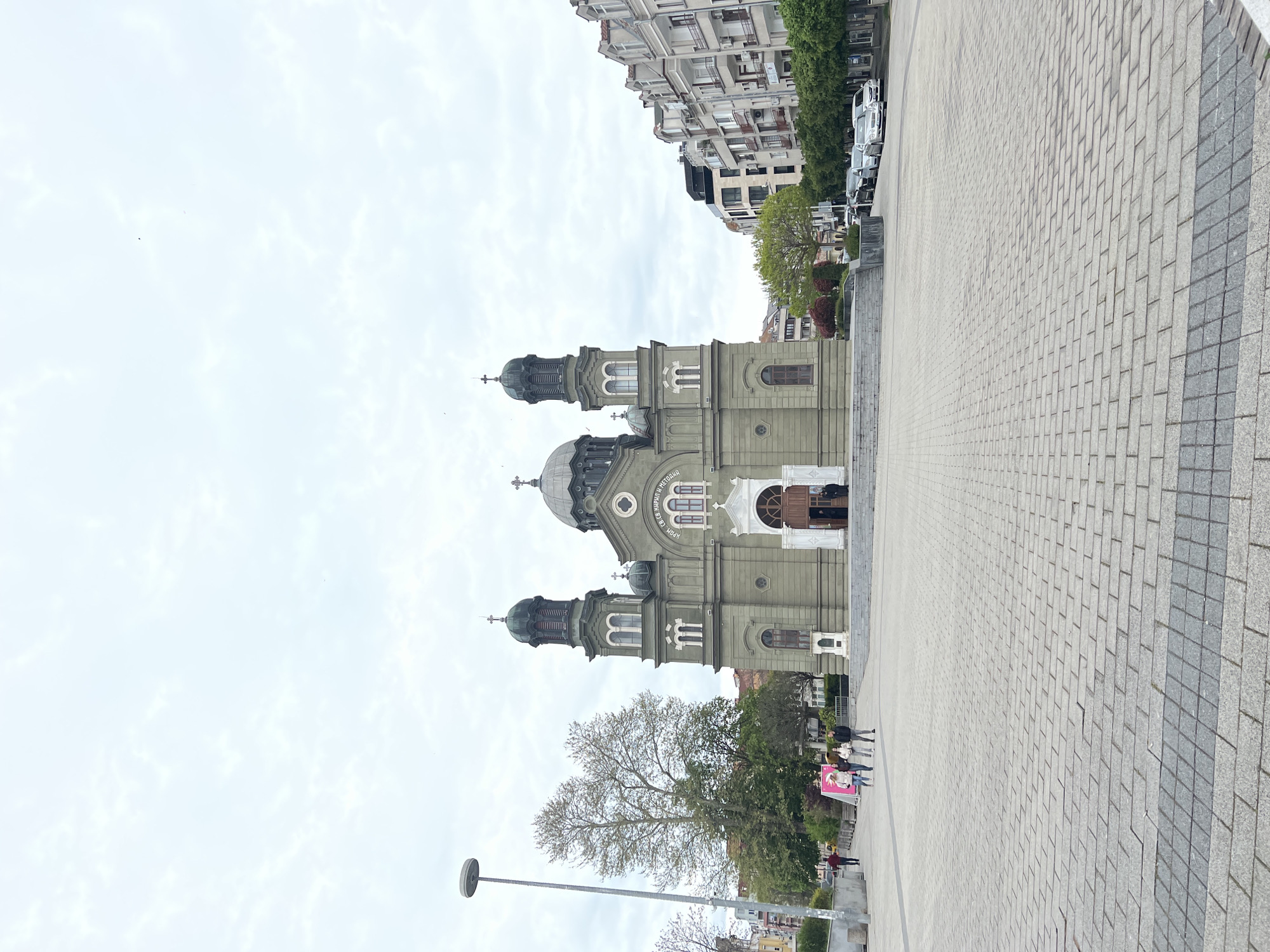 This image captures the majestic Dormition of the Theotokos Church in Burgas, Bulgaria, set against a wide, paved public square under an overcast sky. The prominent church, positioned on the left, showcases a distinctive grey-green facade with multiple domes and bell towers, each topped with an Orthodox cross. Arched windows and intricate architectural details are visible, along with Cyrillic text on its upper section, "УСПЕНИЕ БОГОРОДИЧНО," confirming its dedication to the Dormition of the Theotokos.
The foreground and right side of the composition are dominated by a spacious plaza, paved with light-colored rectangular blocks. A few people are discernible in the distance on the plaza, appearing small and engaged in casual walking or standing near the church. Further back, several multi-story urban buildings with balconies and commercial facades line the street, partially hidden by trees, some with lush green foliage, others still budding or sparsely leafed, suggesting early spring. Parked vehicles are faintly visible alongside these background structures. A contemporary street lamp is positioned in the lower left corner of the frame.
The photograph was taken during daylight hours, characterized by a bright yet uniformly cloudy sky, resulting in soft, shadowless illumination. The scene conveys a tranquil, everyday ambiance in an urban setting. The unique architectural style of the church and the visible Cyrillic script provide strong contextual clues to its location in Burgas, Bulgaria.